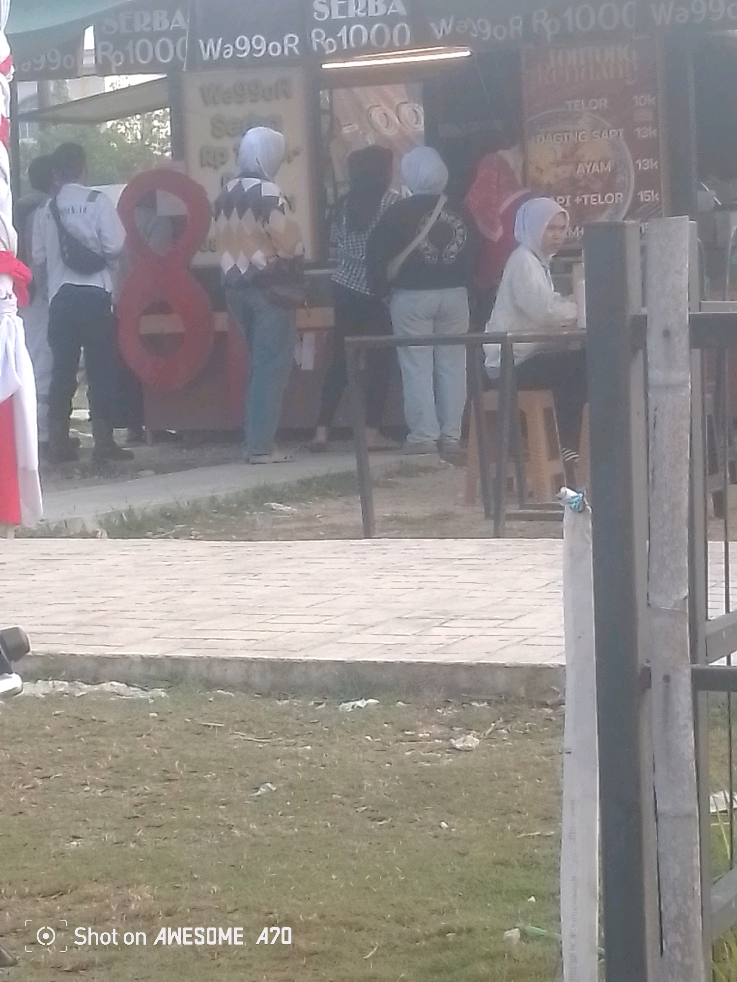 The image depicts an outdoor scene in Batam, Indonesia, likely a street food stall or market area. In the background, a stall is visible with various signs indicating food items and prices. Some signs read "SERBA," "Rp1000," and "Wa99oR," along with prices like "10k," "13k," and "15k." Specific food items mentioned include "TELOR" (egg), "DAGING SAPi" (beef), and "AYAM" (chicken).
Several people are present. On the left, two individuals are standing, one wearing a white shirt with black lettering and a black fanny pack, and another in a white hijab and a patterned sweater. To their right, a person in a white hijab and sweater is visible, and another person in a white hijab is seated on a stool. Further to the right, a person in a red garment is standing.
In the foreground, a red, abstract shape resembling the number "8" is prominent. The ground is paved with square tiles, and beyond that, there is a grassy area with scattered debris. A concrete pillar and a metal fence are also part of the scene. The overall lighting suggests it is daytime, possibly late afternoon, with a slightly hazy quality to the image. The bottom left corner of the image has a watermark that reads "Shot on AWESOME A70."