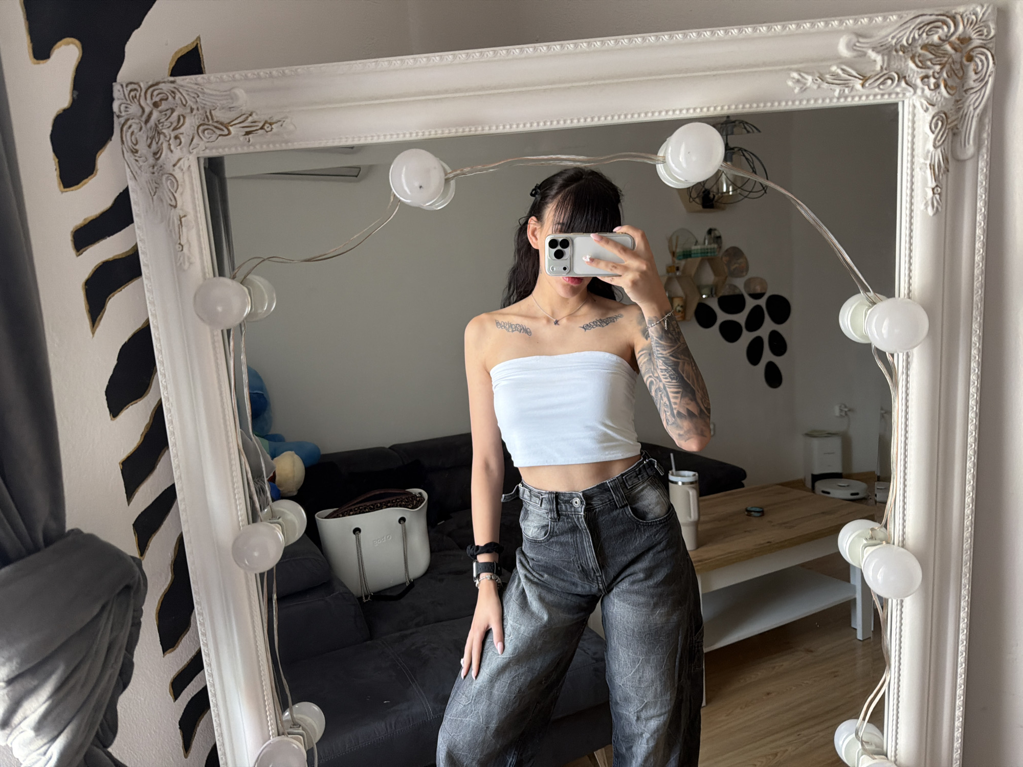 The image shows a young woman taking a selfie in a large, ornate, white-framed mirror. She is wearing a white strapless top and dark, baggy jeans. She has tattoos on her arms and chest. The mirror has lights around its perimeter. In the background, a portion of a dark grey couch, a white bag, and a wooden table with chairs are visible. The room appears to be a bedroom or living area. The image does not provide any specific details about the location being Plovdiv, Bulgaria.