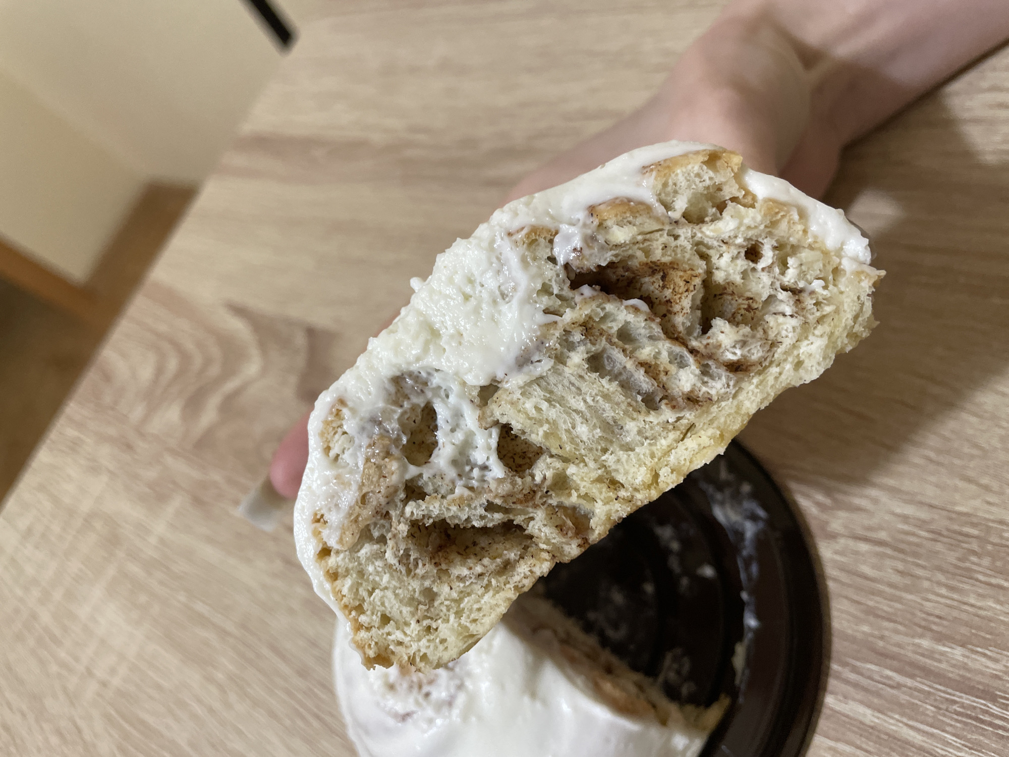 A close-up shot focuses on a cinnamon roll, broken in half to reveal its interior. The roll has a light-colored dough, swirled with cinnamon filling. A thick layer of white icing covers its exterior. The top half of the roll is being held in the air, exhibiting the exposed inner layers. A person's hand is partially visible holding the roll. The bottom half sits on a dark-colored plastic container. The roll and container are set on a light brown wooden surface. The location is Ufa, Russia, though the exact location in the city cannot be determined from the image.