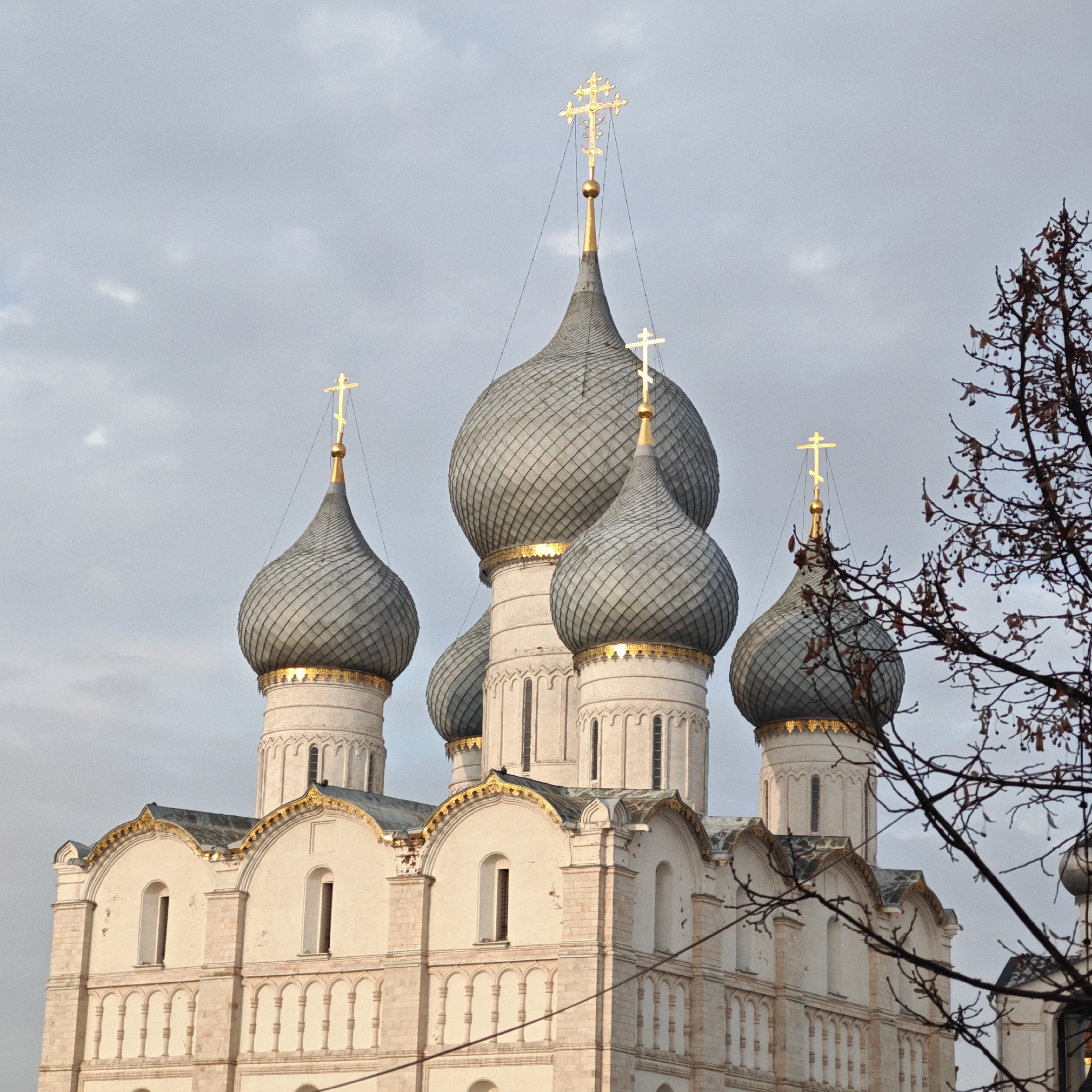 The image depicts the Dormition Cathedral in Rostov, Russia, showcasing its distinctive architecture. The building features white stone walls adorned with arched windows and decorative detailing, topped by a series of bulbous, gray-tiled domes. Golden crosses surmount each dome. The sky is overcast. A bare tree branch partially obscures the lower right corner of the frame. No people are visible in the scene.