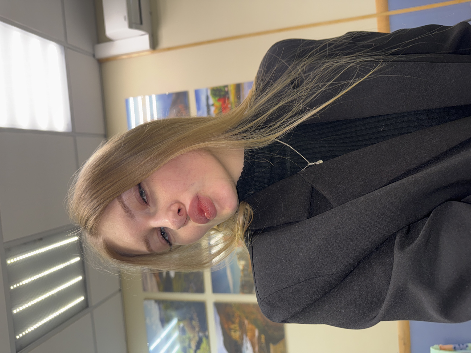 A blonde woman with blue eyes and pursed lips is wearing a black turtleneck sweater and a black blazer. She is indoors, possibly in an office or school setting, as evidenced by the fluorescent lighting and wall art visible in the background. The location is implied to be Perm, Russia due to the prompt's specifications.