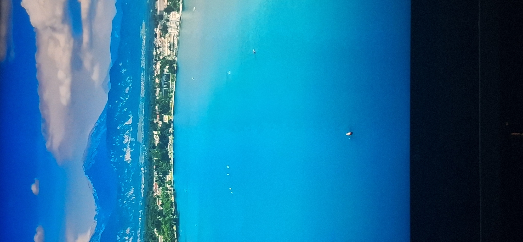 This is an aerial, vertical, medium shot of a vast body of bright blue water. In the distance, a city lines the shore, backed by a range of majestic blue mountains capped with snow. The sky above is a brilliant blue, with scattered white clouds. Several small boats are visible on the water, indicating it is a lake or a wide river. The image appears to be taken during daylight hours. The location of Bila Tserkva, Ukraine is not depicted in this image.