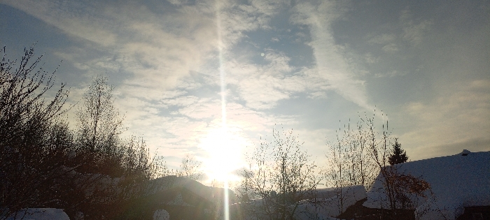 The image depicts an outdoor winter scene in Красный Маяк, Russia, characterized by strong sunlight filtering through an overcast sky. The sun, positioned low in the sky to the left of the central axis, emits a prominent vertical lens flare. The sky above is predominantly light blue with scattered white and grey cloud formations. In the foreground and midground, numerous deciduous trees and shrubs are visible, their bare branches silhouetted against the bright sky. The ground and visible surfaces, including what appears to be a residential rooftop on the far right, are covered with a layer of white snow. Another dark, conical coniferous tree is partially visible behind the bare branches on the right. No human figures, animals, or discernible actions are present in the frame. The overall impression is of a bright, cold, snow-covered environment.