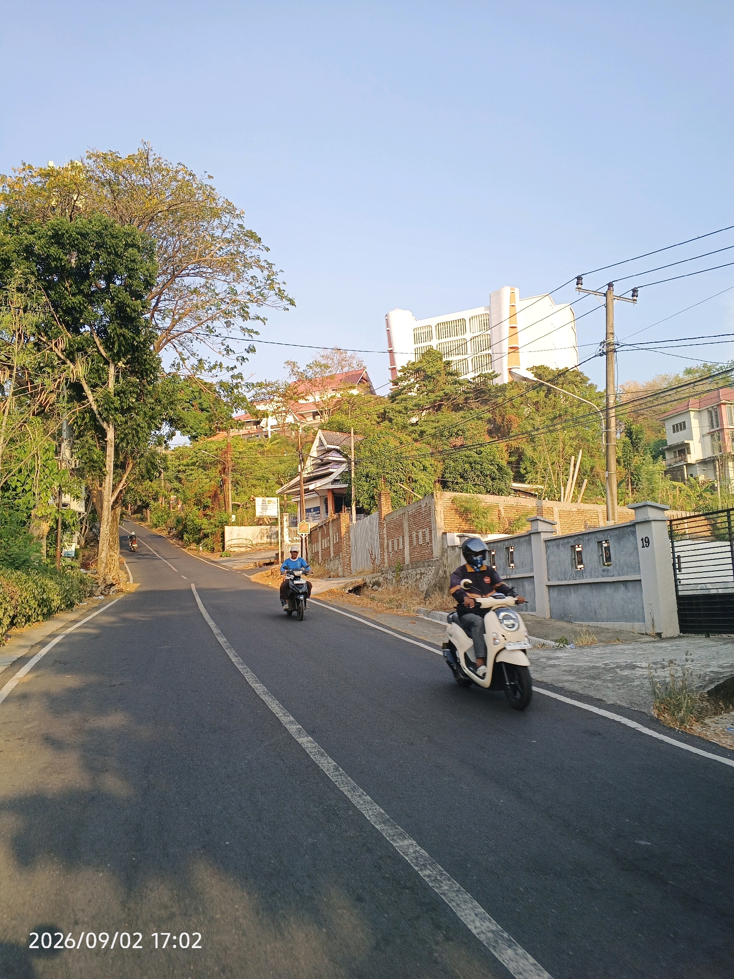 This image depicts a street scene in Pare-Pare, Indonesia, likely during the late afternoon as indicated by the timestamp and the warm lighting. A paved road, marked with a white double line, curves uphill. Two individuals are riding motorcycles on the road. In the foreground, a person on a cream-colored scooter with a helmet is riding towards the viewer's left. Further up the road, another person on a dark-colored motorcycle is also riding uphill.
The environment is characterized by lush green trees and vegetation lining the road. On the right side of the road, a gray fence with a "19" marker is visible, along with a black gate and part of a building. In the background, several multi-story buildings are situated on a hillside, one of which is a large, modern white building with rows of windows. The sky is clear and blue. The overall impression is of a moderately built-up residential or commercial area on a hilly terrain, with a clear sky and warm sunlight casting shadows on the road.