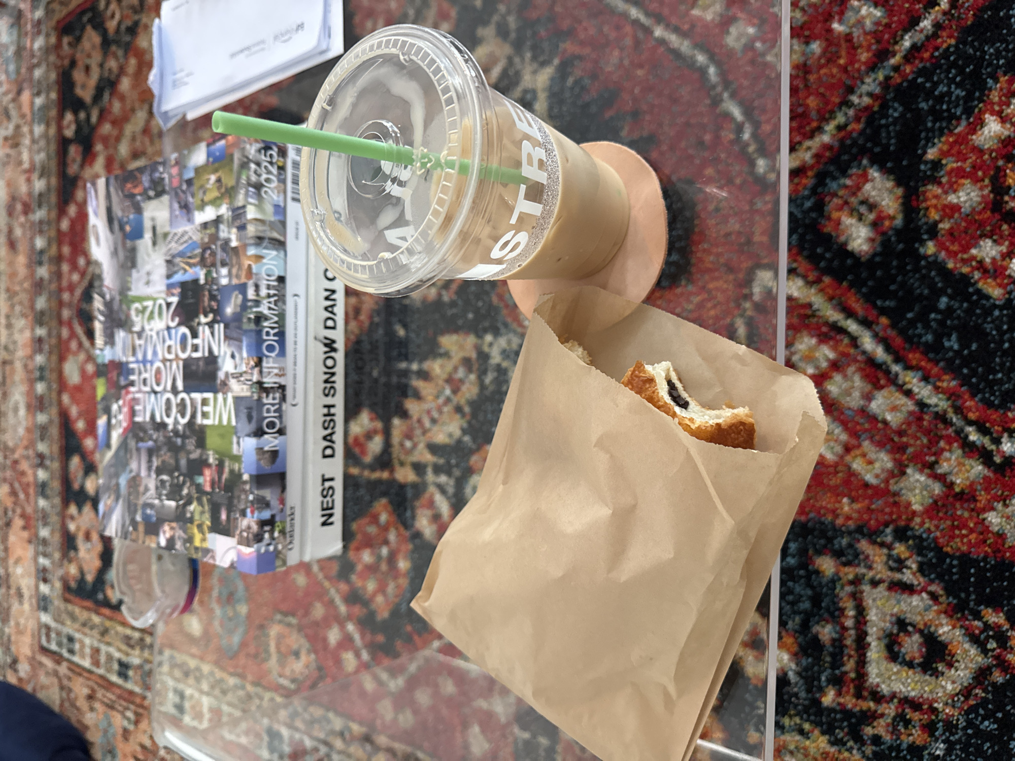 An overhead, slightly tilted shot captures a casual scene on a clear coffee table, set over a richly patterned rug. The table holds a light brown paper bag containing a partially eaten pastry with a dark filling, possibly chocolate. Next to it is a clear plastic cup filled with iced coffee, a green straw protruding from the lid. White, textured letters spelling "STRE" are visible on the side of the cup, suggesting a brand name. The cup rests on a round, light brown coaster.
Beneath the cup and pastry bag, a stack of books or magazines is arranged. The top item features a collage-style cover with various images and the prominent, reversed text "WELCOME MORE INFORMATION 2025". Below it, the spine of another book is visible, reading "NEST DASH SNOW DAN C".
The clear tabletop reflects the intricate designs of the rug underneath, which boasts a vibrant palette of red, black, and lighter gold/white patterns. The overall impression is an indoor setting, perhaps a living room or a relaxed lounge area, where someone is enjoying a coffee and snack, possibly while reading. The time of day and weather are not discernible. No people are visible in the image.
