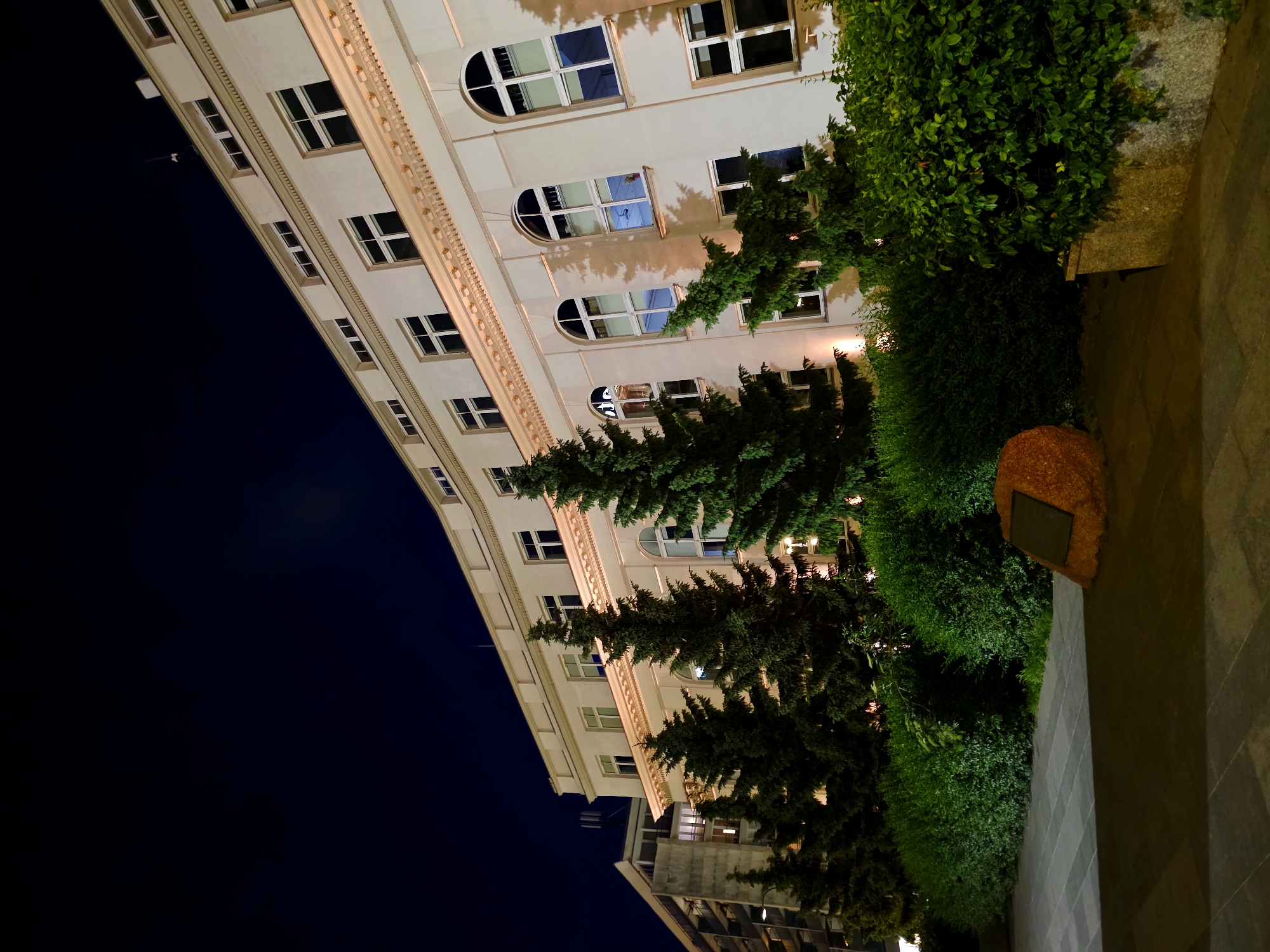 The image depicts a building at night in Warsaw, Poland. The building is a multi-story structure with a white facade, featuring a row of windows on each level. The architecture suggests it could be an institutional building or a historic residence. Several tall, dark green coniferous trees stand in front of the building, partially obscuring the lower floors. A neatly trimmed hedge runs along the base of the trees. In the foreground, a paved pathway is visible, leading towards a large, reddish-brown rock with a plaque attached to it. The sky is dark, indicating nighttime, and the building is illuminated by artificial lights, casting shadows on its facade. To the left of the main building, another structure with a more modern architectural style is partially visible, also lit by artificial lights. The scene is calm and still, with no people or activity evident.