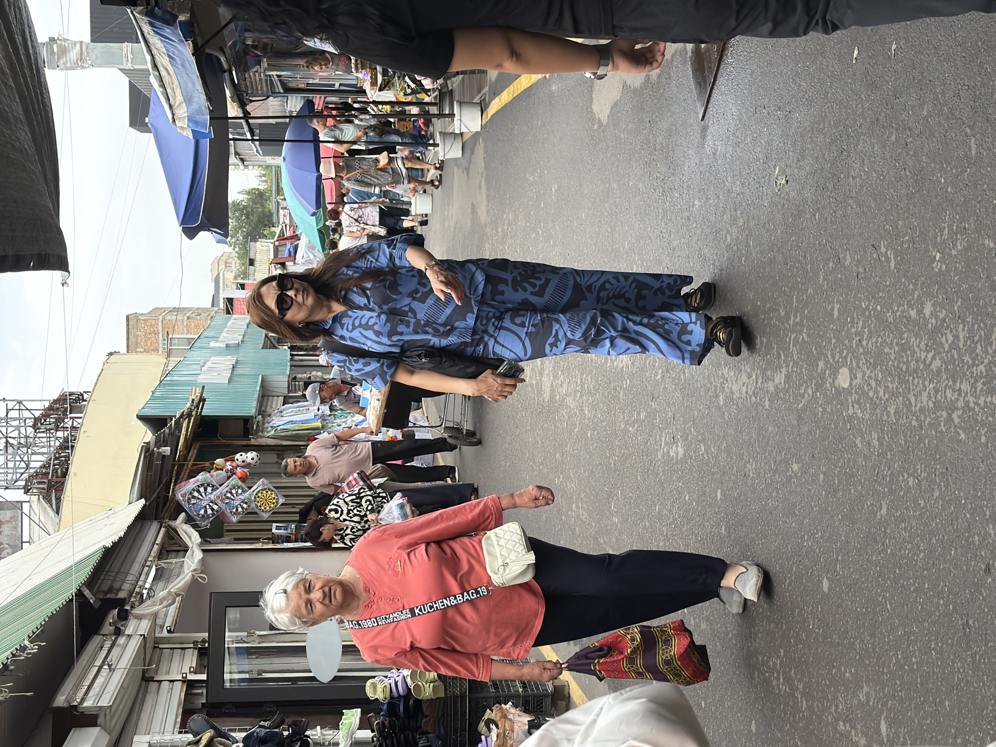 An outdoor market scene unfolds with two women walking towards the viewer. The woman on the left, an older Caucasian female with gray hair, wears a salmon-colored, button-down shirt and dark pants, carrying a patterned shopping bag in her left hand and a small white purse over her right shoulder. The woman on the right, a middle-aged Asian female with dark hair, sports sunglasses and a blue patterned pantsuit, with a black bag slung over her shoulder. They walk on a paved street lined with market stalls and awnings. Various people and merchandise are visible in the background. The environment appears to be an urban market setting.