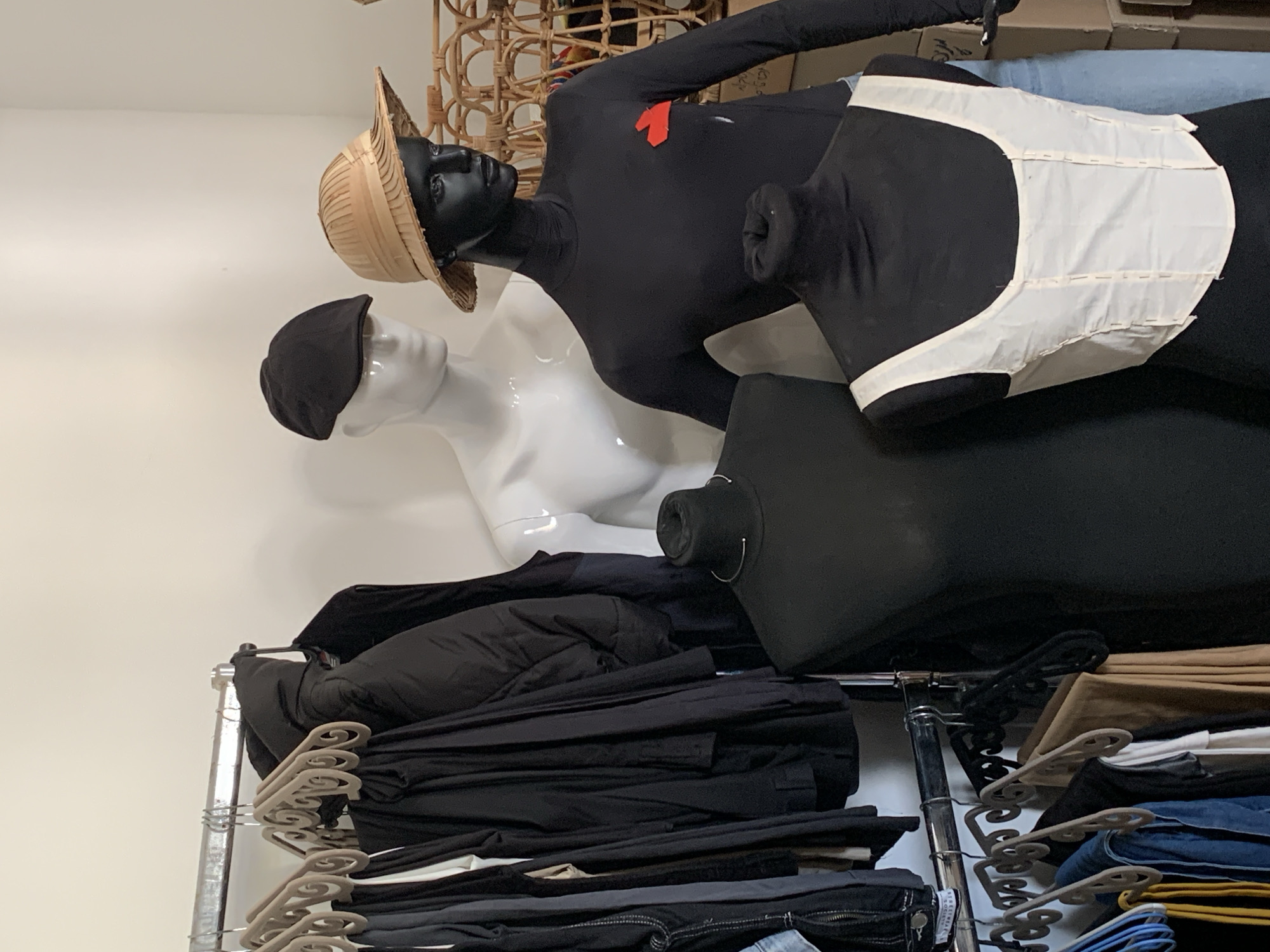 The image displays an indoor retail environment, likely a clothing boutique, with light-colored walls. No specific location within Tel-Aviv is identifiable from the visual information.
The scene features two chrome clothing racks on the left. The upper rack holds numerous dark-colored garments, predominantly black jackets and trousers, suspended on light brown velvet hangers and some black plastic hangers. The lower, partially visible rack displays additional clothing items, including denim and tan trousers, on various hangers.
Four mannequins are present:
1. A white, male-presenting mannequin, wearing a black flat cap, is positioned behind the upper clothing rack, partially obscured.
2. A black, female-presenting mannequin stands to the right, wearing a wide-brimmed straw hat, a long-sleeved black turtleneck shirt with a red fabric appliqué on the left chest, and light blue jeans.
3. In the immediate foreground, a black torso display form wears a silver-colored choker.
4. Adjacent to it, another black torso display form exhibits a white sleeveless top with visible vertical stitching or pins, resembling a corset.
In the background, to the right of the black female mannequin, a light brown woven rattan object, possibly a basket or furniture piece, is partially visible. Stacked cardboard boxes are also partially visible on the far right, suggesting inventory or storage.
