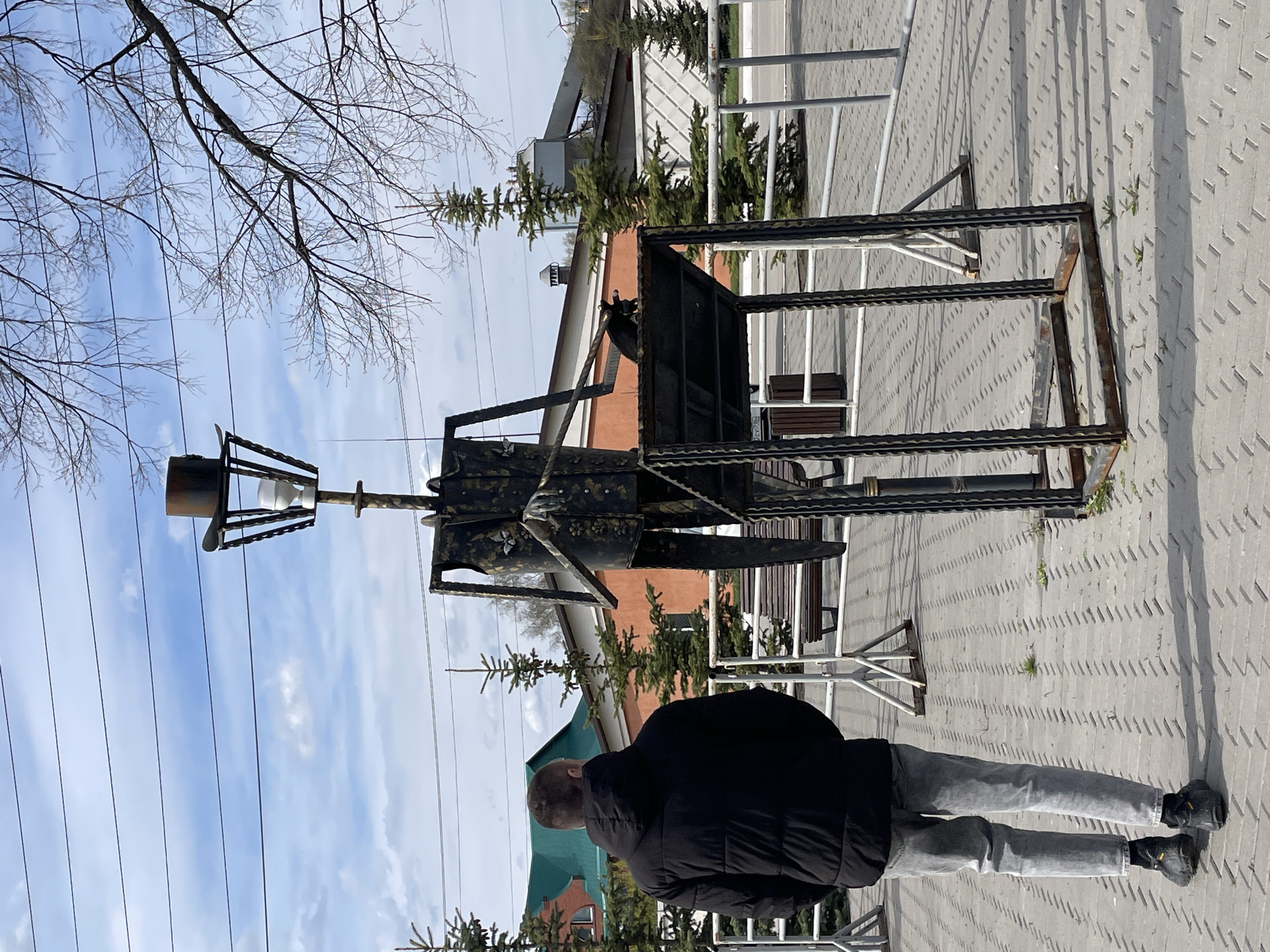 A man stands facing a metal sculpture in Urussu, Russia. The sculpture appears to be an abstract representation of a person or figure, constructed from metal rods and plates. The sculpture features a head-like structure with a cylindrical object at the top. It is standing on a paved surface with a patterned texture. Behind the sculpture are benches and some evergreen trees, with a building visible in the background. Overhead, there are tree branches and power lines against a partly cloudy sky. The man is wearing a dark jacket and gray pants and is looking at the sculpture.