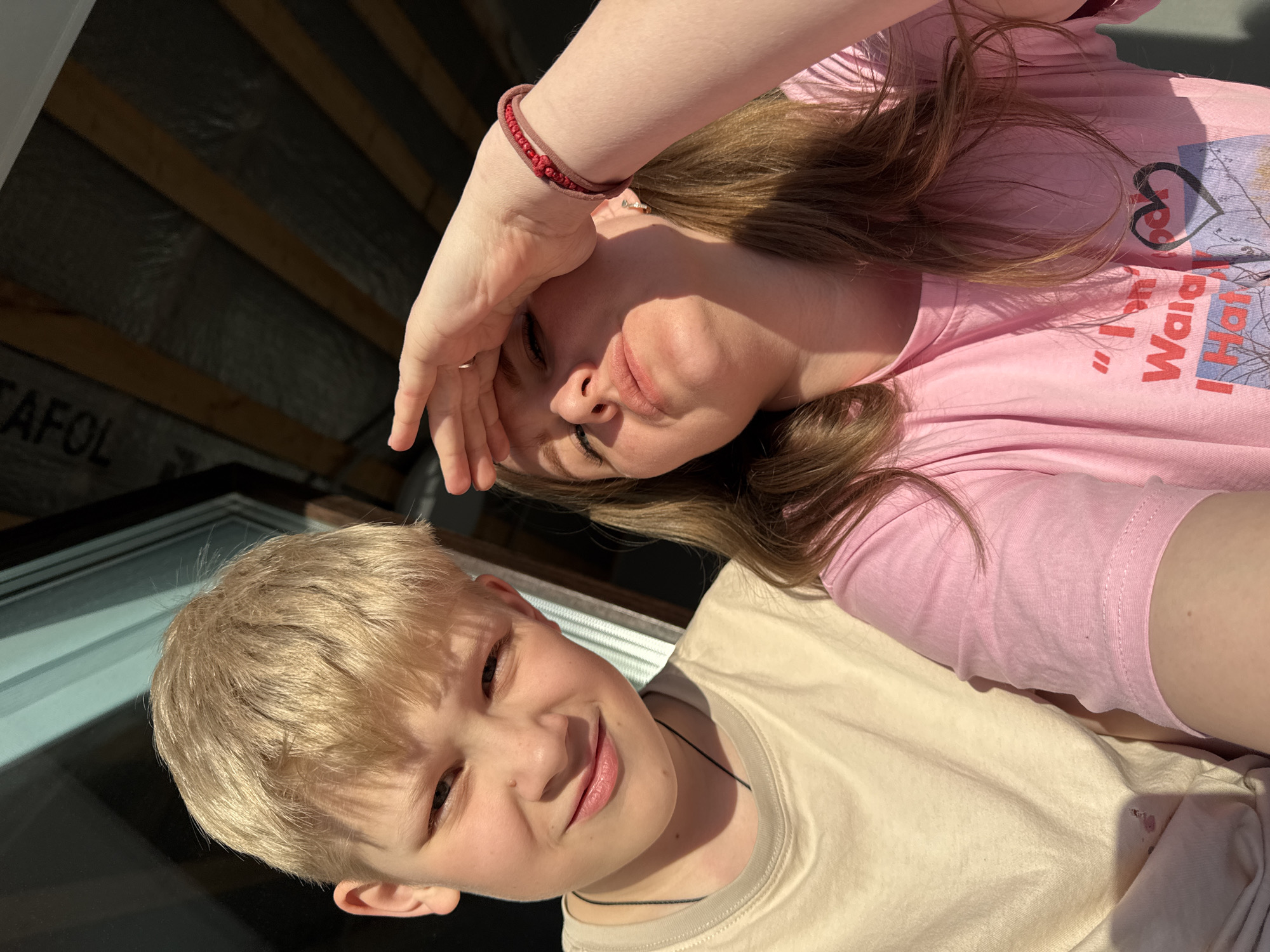 The image displays two individuals in Gornyi (Dvurechenskoe S/P), Russia, captured under direct sunlight. On the left, a male child with short blonde hair, fair skin, and blue eyes looks forward with a slight smile, wearing a light beige t-shirt and a thin dark necklace. On the right, an adult female with light brown hair and fair skin looks upward, shielding her eyes from the sun with her raised right hand. She wears a pink t-shirt with a partially visible graphic print, a red braided bracelet on her right wrist, and a gold-colored ring on her right ring finger. The background features a reflective surface on the upper left and an overhead structure of dark wooden beams supporting a grayish material, with the text "AFOL" partially visible on the upper right.