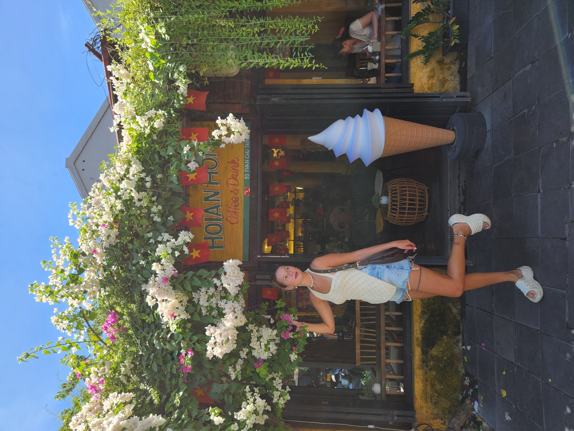 A young woman stands in front of a cafe in Hoi An, Vietnam. She is wearing a white crochet tank top, denim shorts, and white platform sandals. Her hair is pulled back in a ponytail, and she is accessorized with a beaded necklace and a brown crossbody bag. She is posing with one leg extended. The cafe, named "HOIAN HOMIES Coffee & Drink," has a wooden facade decorated with red Vietnamese flags and cascading greenery. A large, illuminated ice cream cone sculpture stands outside the cafe entrance. Inside, a woman is seated at a table. The street in front of the cafe is paved with dark tiles. The sky is clear and blue.