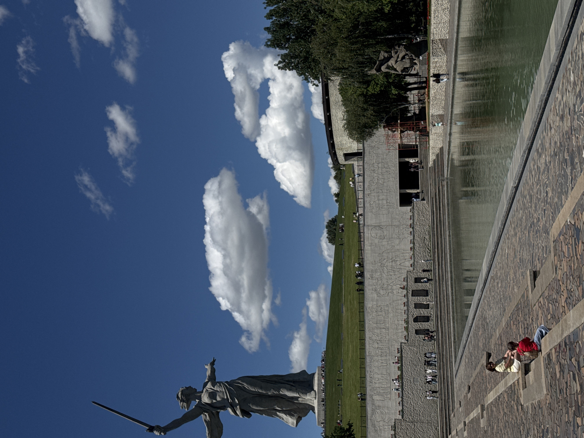 The image shows the "The Motherland Calls" statue in Volgograd, Russia, part of the Mamayev Kurgan memorial complex commemorating the Battle of Stalingrad. The statue is prominently displayed against a blue sky with scattered clouds. In the background, a stepped memorial complex ascends a hill, with a long, rectangular reflecting pool running alongside. People are visible on the steps and the grassy slopes of the memorial. A few individuals are sitting on a wall in the foreground. The complex is surrounded by trees.