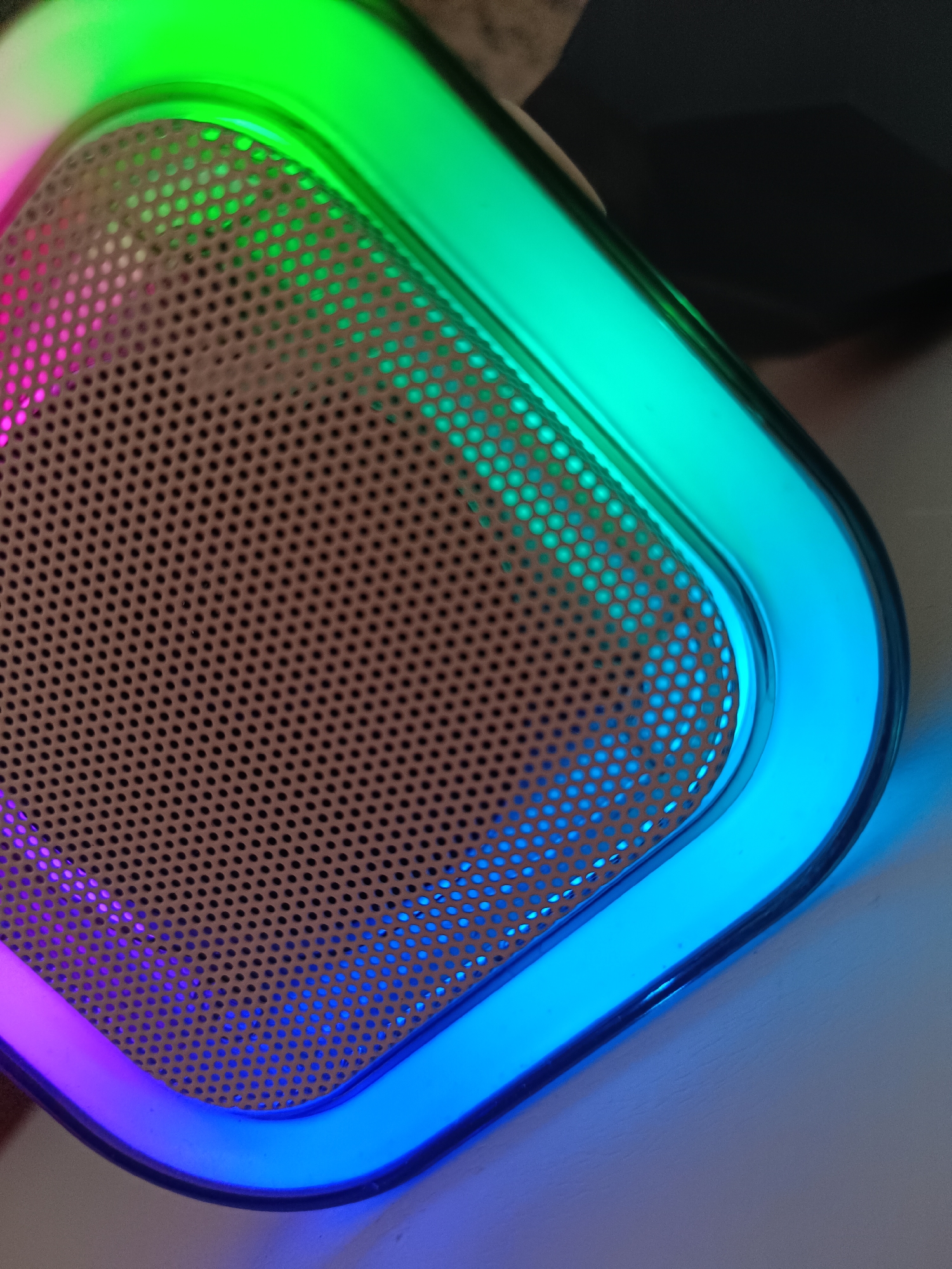 A close-up shot shows a corner of a device, likely a speaker, with a perforated metal grill. The frame surrounding the grill is illuminated with a color gradient ranging from green to blue to purple. The background is out of focus. The lighting is artificial. There is no indication of Tobolsk, Russia, or any location in the image. No people, subjects, actions or interactions are visible.