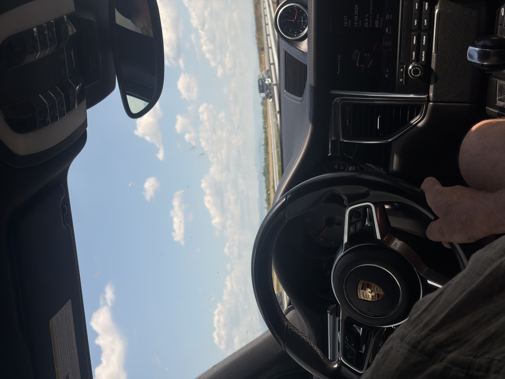 The image shows the interior of a Porsche, likely a Macan, from the perspective of the driver. The driver's hands are on the steering wheel, and part of their arm and leg are visible. The dashboard features a Porsche emblem on the steering wheel and a digital display showing trip information, time, date, outside temperature (29.0°C), and range (465 km). A clock on the dashboard shows 12:17. The exterior view through the windshield reveals a highway with other vehicles in the distance and a bright, cloudy sky. The location is identified as Загорци, Bulgaria.