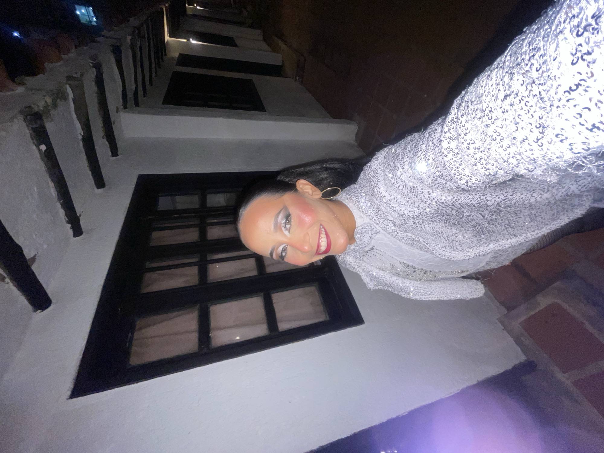 The image shows a person in a sequined jacket and white top posing for a selfie outdoors at night. The background features a white wall with dark wooden beams, a black-framed window, and a brick pathway. The location is identified as being in the city of Porlamar, Venezuela. The person has dramatic makeup with bright eyeshadow, blush, and lipstick, and their hair is pulled back in a ponytail. They are wearing large hoop earrings. The lighting is dim, with some artificial lights visible in the background, creating a slight purple hue on the left side of the image.