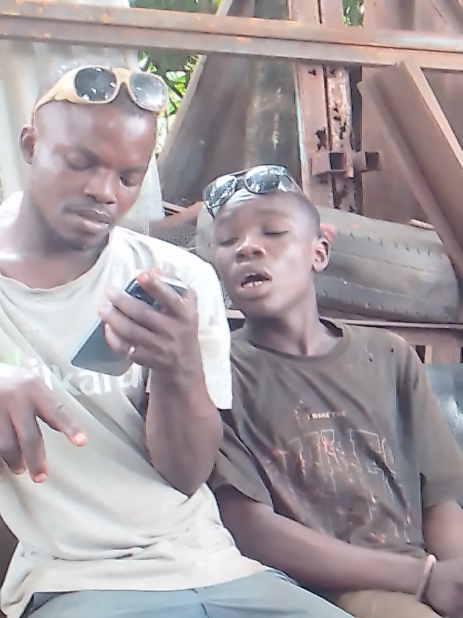 The image features two males, appearing to be of African descent, seated in what looks like an outdoor or semi-outdoor industrial setting during the daytime.
The subject on the left is an older male with light brown skin and a shaved head, on which he wears a pair of light-colored sunglasses. He is wearing a light-toned, possibly off-white, short-sleeved t-shirt. He is engrossed in a black smartphone which he holds with both hands, his gaze directed downwards at its screen. Part of a word, "inkan", is visible on his t-shirt. His right hand has a ring on the ring finger, and his left hand is visible with fingers pointing downwards, though the exact context of this gesture is unclear.
To his right sits a younger male, also with a shaved head and sunglasses perched on top. He wears a dark, possibly brown or olive green, short-sleeved t-shirt that shows signs of wear, including visible holes or distressed areas. His mouth is slightly open, and he is looking intently towards the smartphone held by the older male, suggesting he is observing or participating in the activity on the device. Partial text "I WANT TWE..." is visible on his shirt.
The background is characterized by various rust-colored metal structures, possibly beams or scaffolding, indicating an industrial or construction-related environment. A dark, rounded object resembling a tire is visible behind the younger male. Glimpses of greenery through the background structures suggest an open-air setting. The overall impression is casual, with both individuals focused on the shared interaction with the smartphone.