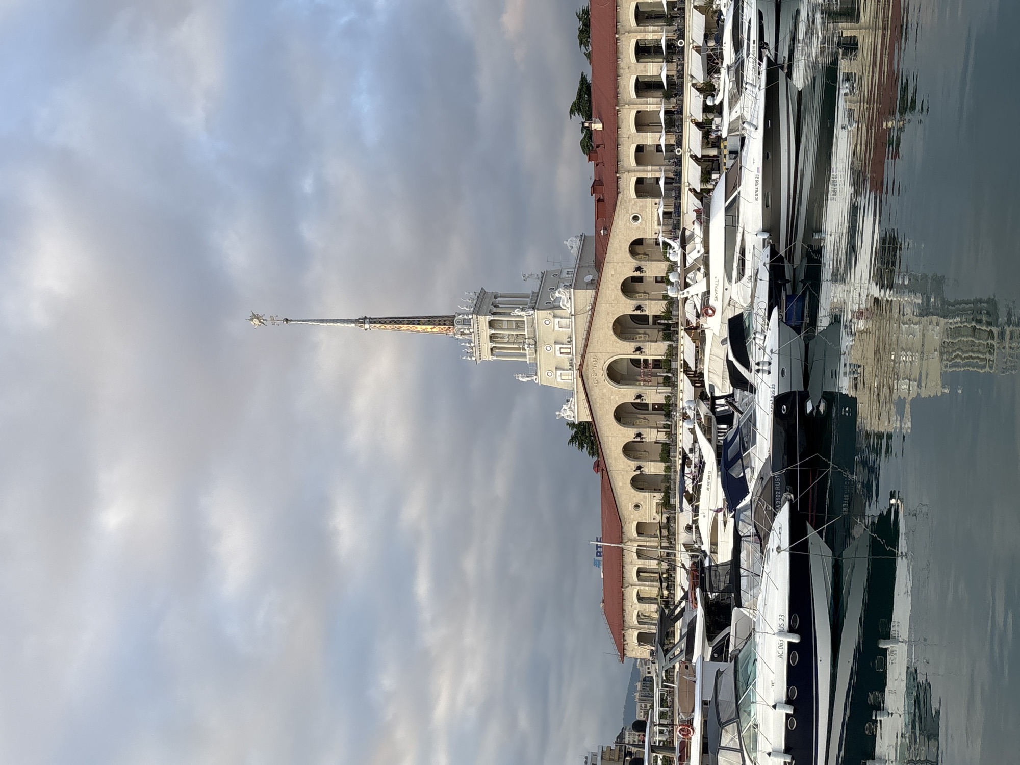 This media file shows a view of a marina in Sochi, Russia. The main subjects are several yachts docked in the water, with a large, ornate building in the background. The building features classical architecture with arched windows, a red tiled roof, and a prominent spire topped with a golden star. The yachts are a mix of white and dark colors, and some have visible registration numbers like "AC 063 US 23," "AA3102 RUS1," and "AC0764 RUS 23." One yacht is labeled "SKYFALL."
The scene appears to be a bright, possibly late afternoon or early morning, given the quality of light and the reflective nature of the water. The sky is cloudy, with soft, diffused light filtering through. The water is calm and reflects the building and yachts, creating a mirror-like effect. There are no people prominently visible in the image. The overall mood is one of tranquility and stillness, characteristic of a harbor. The location is clearly identified as Sochi, Russia, through the provided context. The text visible on the yachts provides specific identifiers for the vessels.