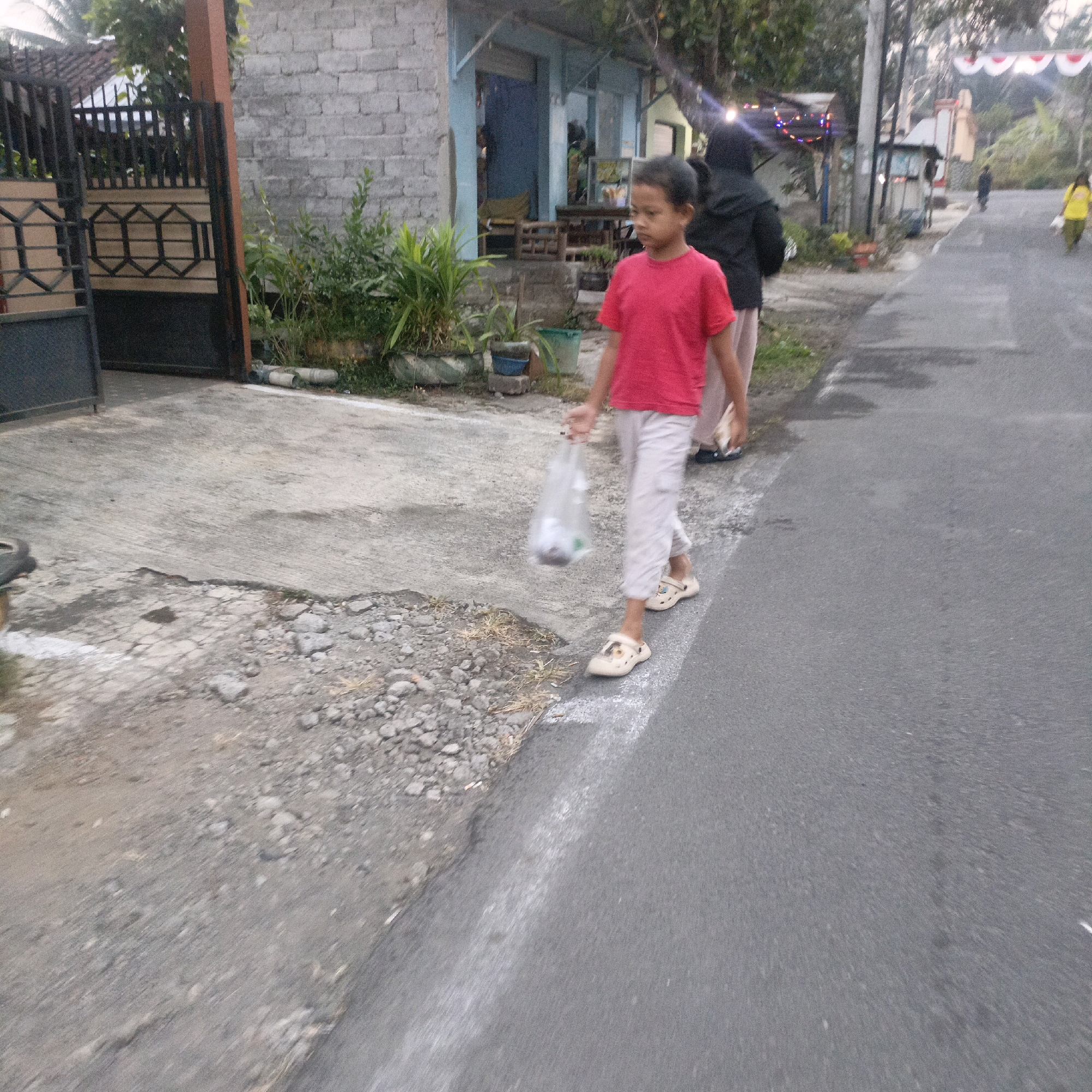 The image shows a young boy in a red t-shirt and light-colored pants walking on a paved road, holding a clear plastic bag. He is wearing sandals. Behind him, a person wearing a black hijab and dark pants is also walking on the road. In the background, there are buildings, some with brick walls, and vegetation. The scene appears to be in a residential or commercial area. The presence of red and white festive banners hanging across the road suggests a possible celebration or event. The overall lighting suggests it is either early morning or late afternoon. This scene is consistent with a street in the city of Salatiga, Indonesia, where such mixed residential and commercial areas, along with common celebrations, are prevalent.