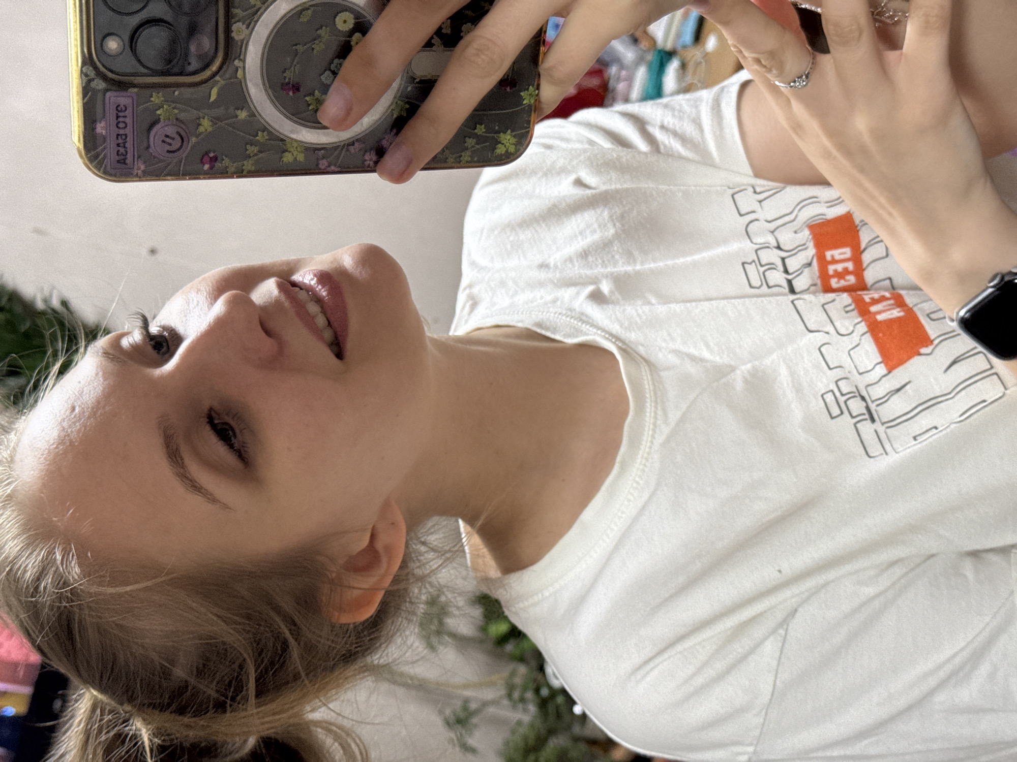 A young, light-skinned woman is taking a selfie, angled to the left. She is holding a phone with a floral-patterned case. The phone's camera array is visible. The woman is wearing a white t-shirt with text in white and orange. She has a ring on her finger and a smartwatch on her wrist. The background is out of focus and includes plants. There are no discernible landmarks indicating the location is Izhevsk, Russia.