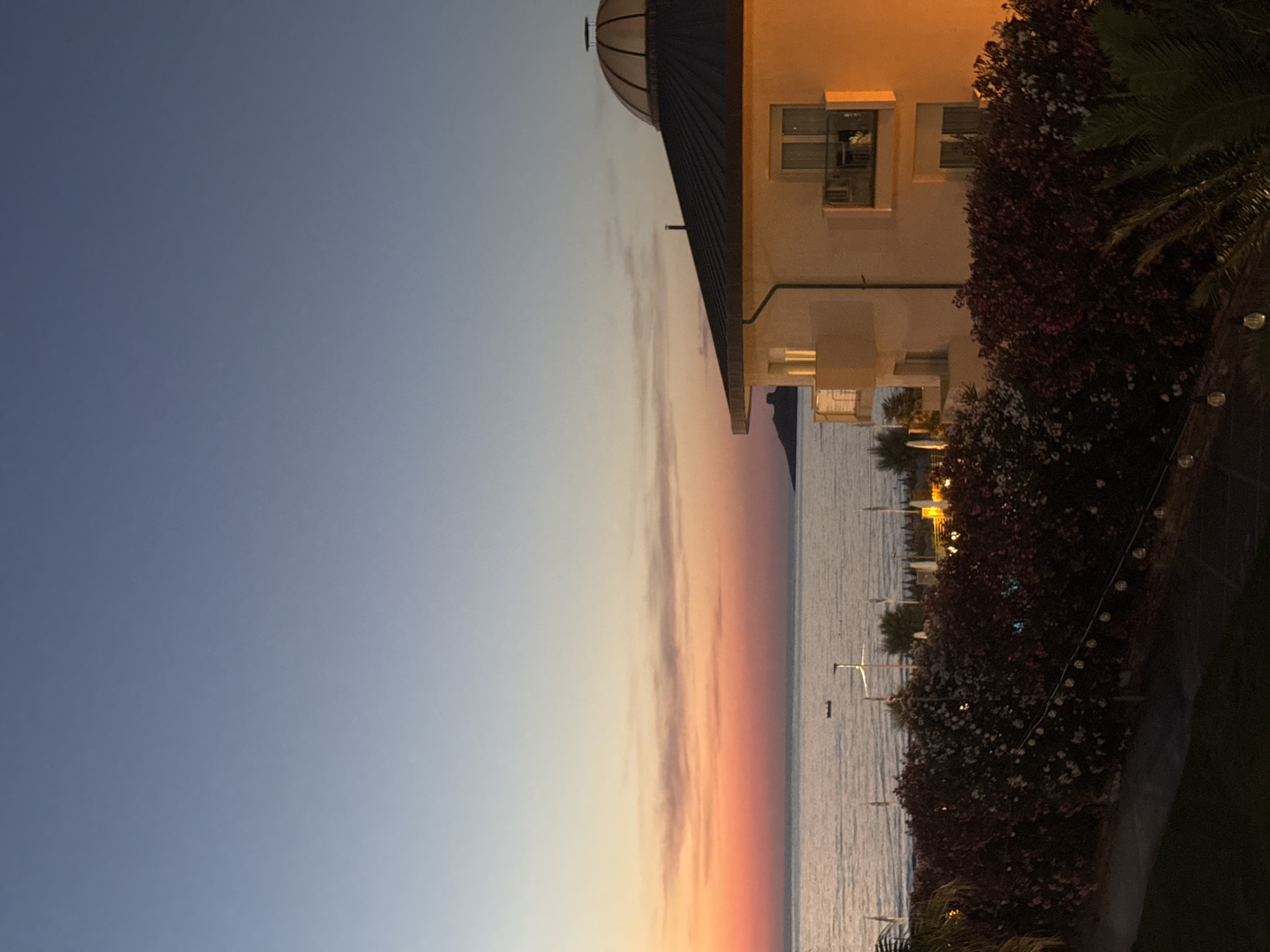 The image depicts a twilight scene at Isola delle Femmine, Italy, overlooking the Mediterranean Sea. A building with a domed roof structure is partially visible on the right side of the frame, illuminated by artificial lights. The building appears to be modern, with visible windows and architectural details. In front of the building, a dense hedge or bush with small red flowers is in bloom, bordering a walkway or path. String lights are hung along the path, suggesting an evening ambiance. The sea is calm, reflecting the soft hues of the sunset. The sky transitions from a clear blue at the top to warm orange and pink tones near the horizon, indicating sunset. The overall mood is serene and picturesque, capturing the beauty of the Italian coastline at dusk. There is no visible text in the image.