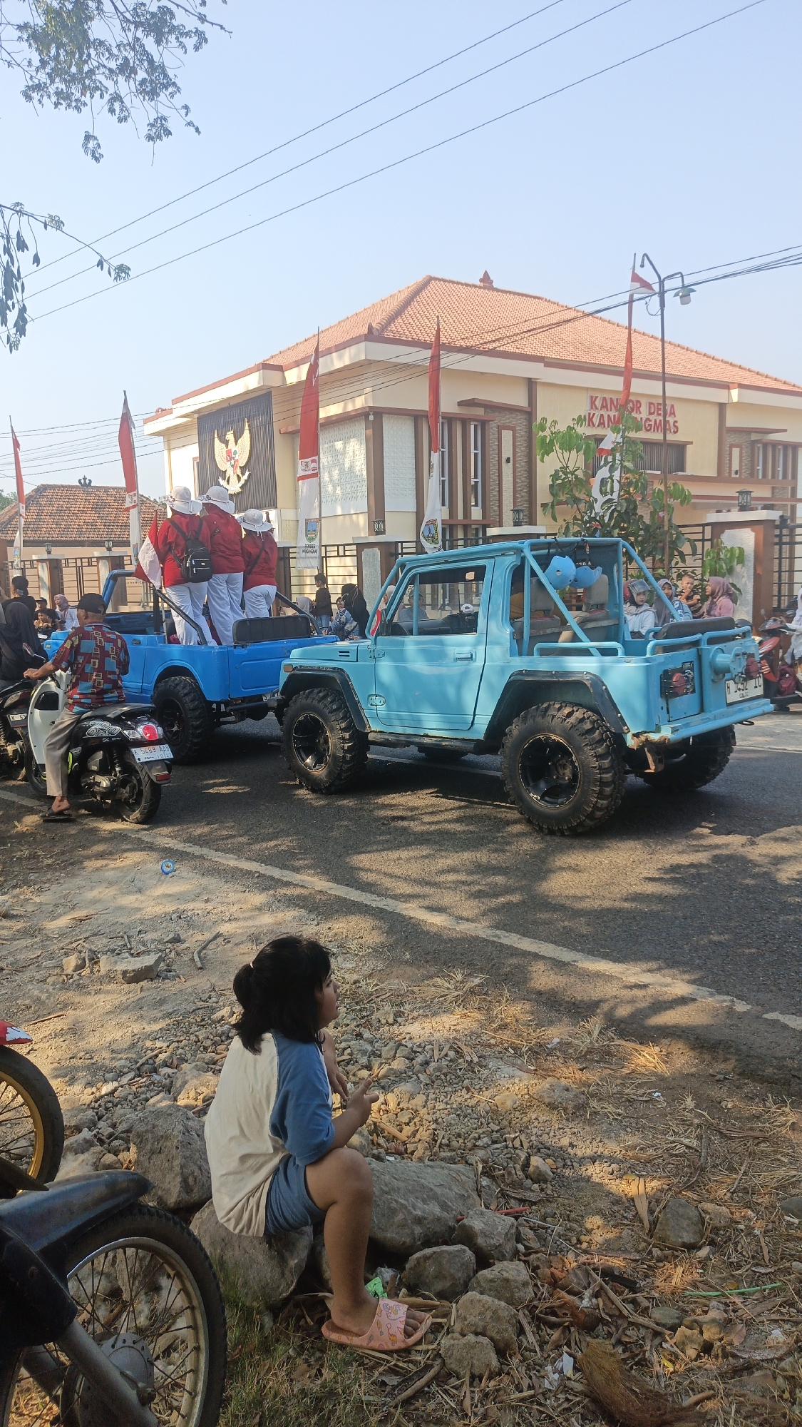 In Kandangmas, Indonesia, a parade features two light blue off-road vehicles driving down a paved road under a clear sky. The first vehicle, a pickup truck, tows the second, a convertible with a roll cage. Both vehicles have large, off-road tires. Several people wearing white hats and red shirts are seated in the back of the pickup truck, holding flags. A young girl with dark hair, wearing a blue and white shirt and shorts, sits on rocks on the side of the road, watching the parade. Other people and motorcycles are visible in the background, and a building with a red tile roof and "KANTOR DESA KANDANGMAS" signage is situated behind the parade. The area is lined with trees and flags.