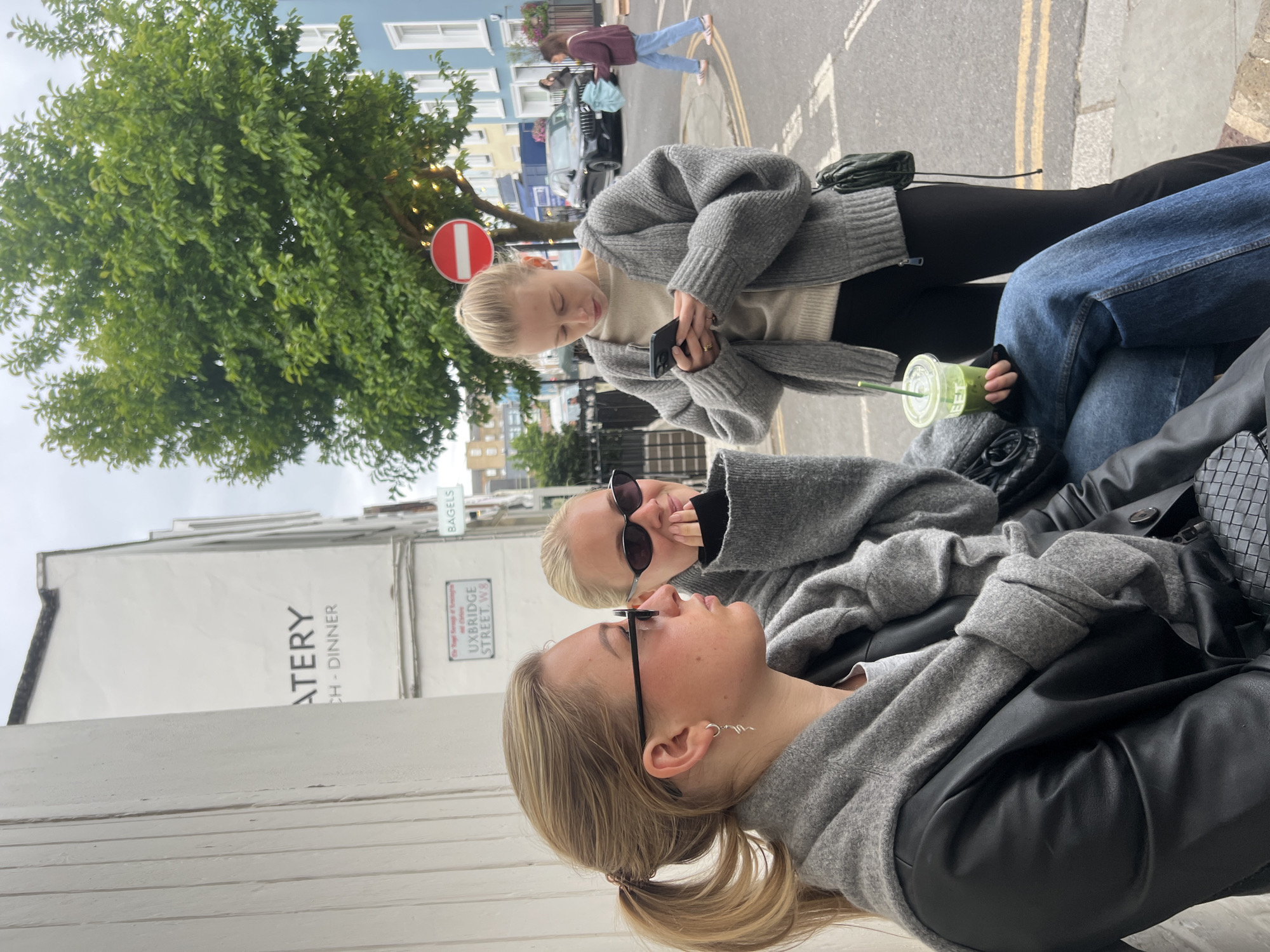 The image shows three young women seated outside a building. One is looking at a phone, the other two are interacting, one wearing sunglasses. All three are wearing gray sweaters. One woman is wearing glasses. A green smoothie sits on the ground. The building signage partially visible reads "ATERY CH - DINNER" and "UXBRIDGE STREET W8". A street sign is visible above the building signage. The location is Uxbridge Street in London. A no entry traffic sign is visible behind the woman with the phone, partially obscured by a tree. Pedestrians and cars are visible in the background. The sky is overcast.
