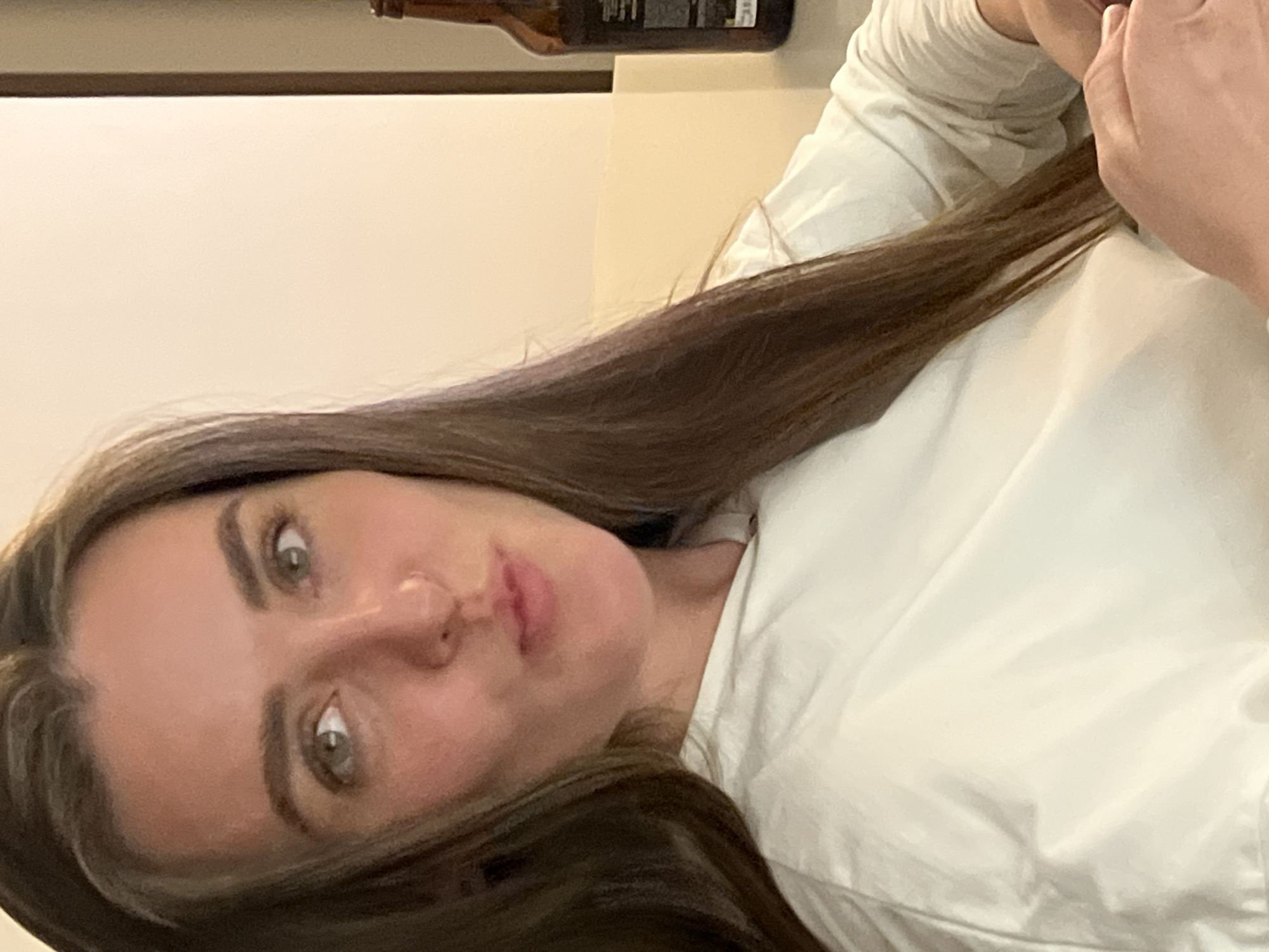 The image shows a woman with long brown hair, wearing a white top. Her face is visible, and she appears to be looking at the camera. The background is blurred and shows what appears to be a shelf with a bottle on it, and an off-white wall. There is no information in the image that indicates a location in the city of Рубцово, Russia.