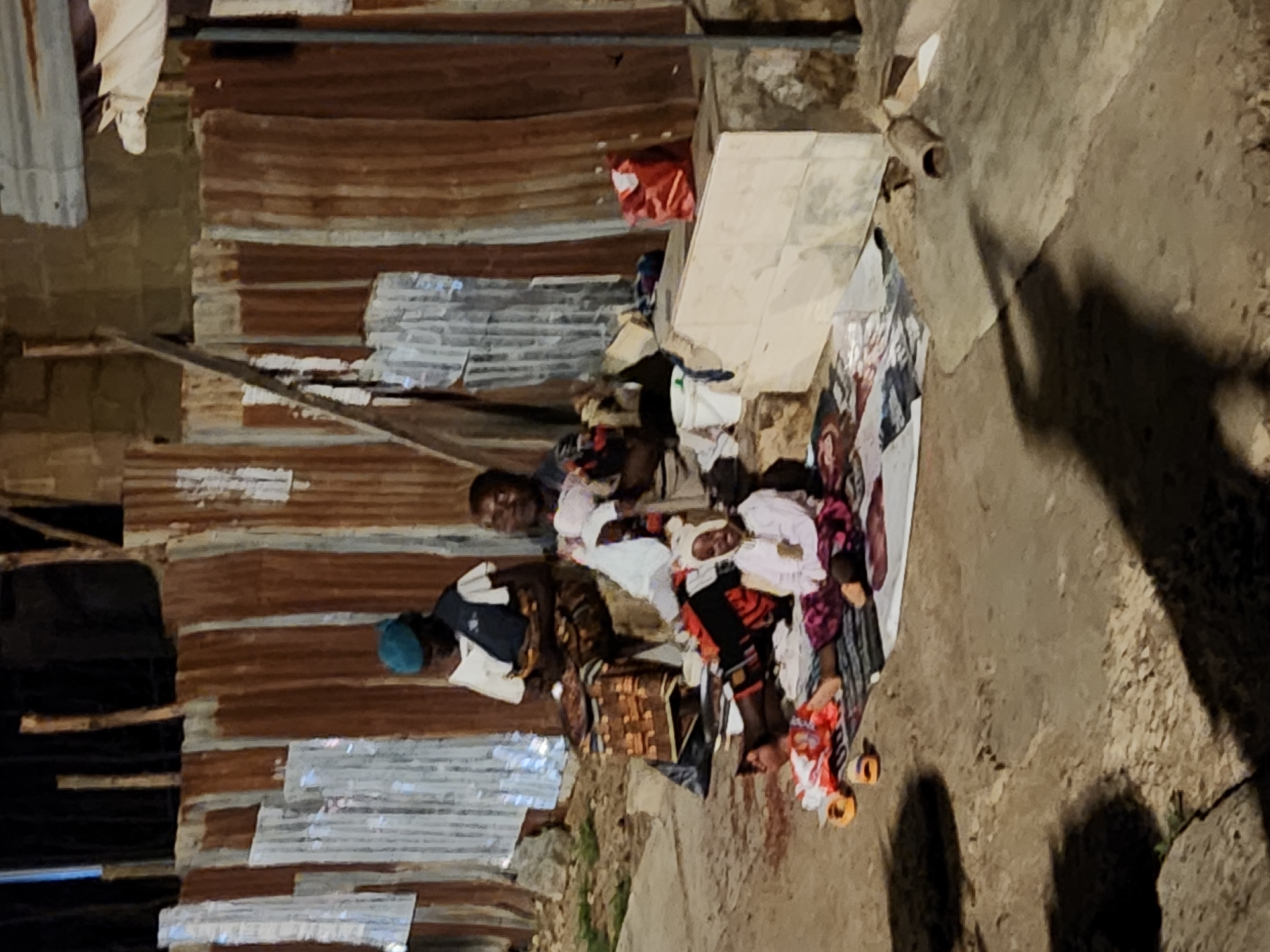 The image depicts a group of people gathered outdoors in Jos, Nigeria, at night. They are positioned on a patterned mat placed on a concrete surface next to a structure made of corrugated metal and brick. The individuals appear to be interacting, with some sitting and others possibly lying down. Various objects such as a bucket, a woven basket, and footwear are scattered around the mat. The environment suggests an informal setting within a populated area.