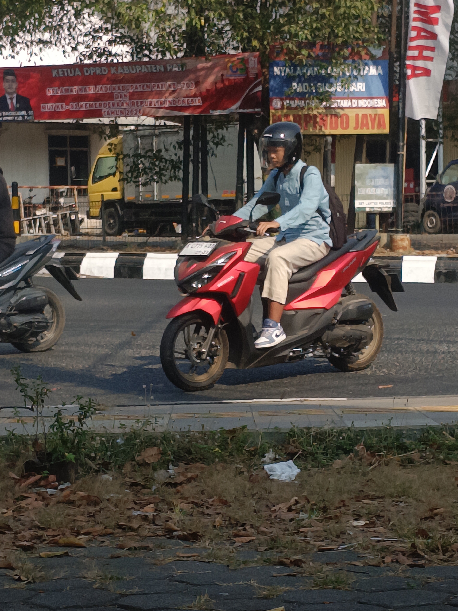 An eye-level, medium shot captures a man riding a red motorcycle on a city street in Indonesia. The man, wearing a light blue long-sleeved shirt, beige pants, a black helmet with a clear visor, and white and blue sneakers, is positioned in the center of the frame. He is looking forward, and his hands are on the handlebars. A dark backpack is visible on his back. The motorcycle is a red scooter-type model, and it is angled slightly to the left.
In the background, several objects and signs are visible. To the left of the motorcycle, another black scooter is parked. Behind the rider, a yellow truck is stopped, and a white banner is draped across a building. The banner has red text and an image of a man. To the right of the rider, a blue and yellow sign with white text is mounted on a pole. Further to the right, a white vertical banner with red text is visible. The street is paved, and a grassy area with fallen leaves lines the side of the road. The sky is overcast with dappled sunlight filtering through trees. The overall scene depicts a typical day on a busy street in an Indonesian city.