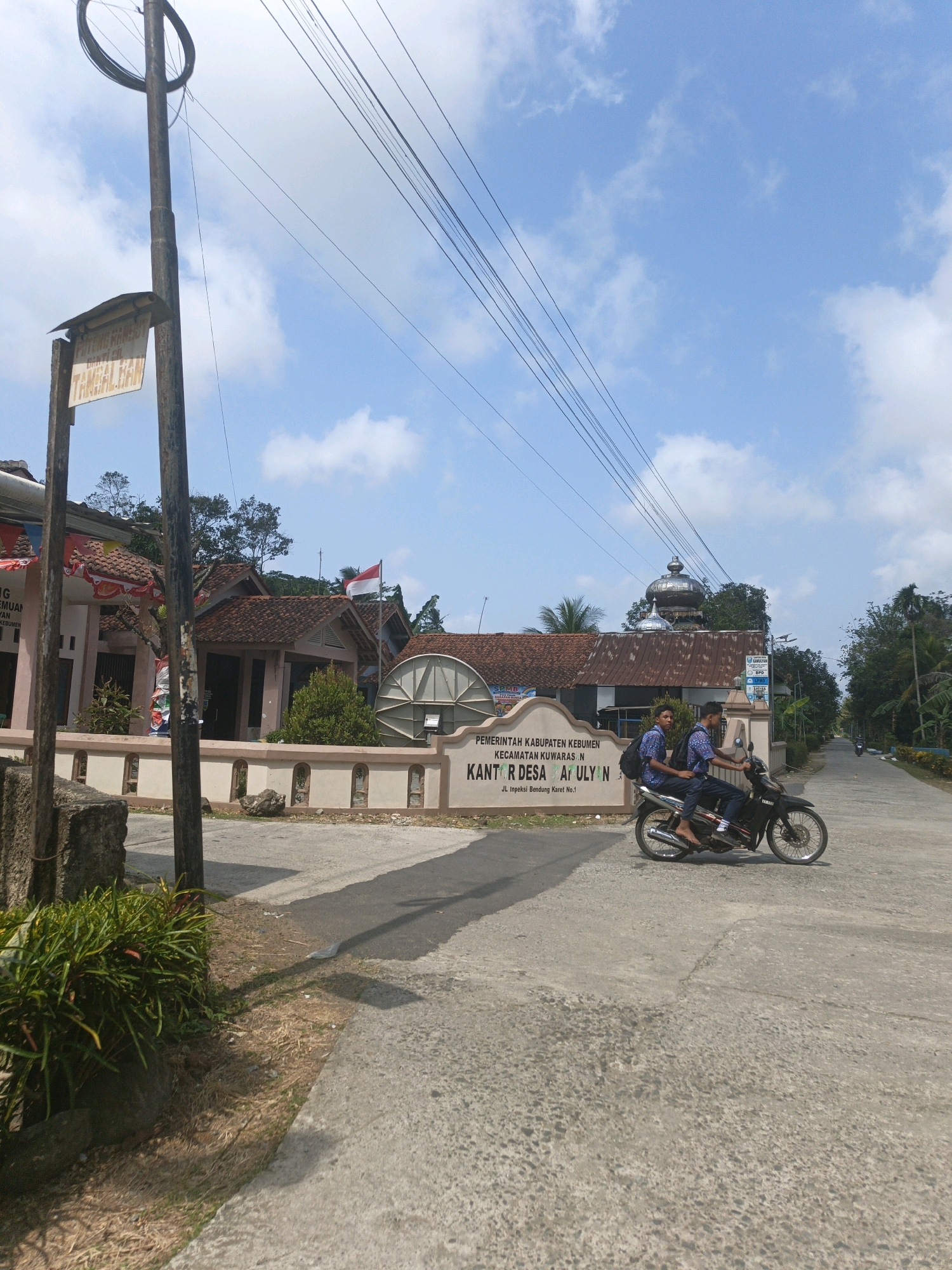 In Kamulyan, Indonesia, on a clear day with scattered clouds, a motorcycle carrying two young men wearing identical blue and white patterned shirts with backpacks rides down a paved road. The motorcycle is black and has a silver exhaust pipe. The men appear to be students. The road passes in front of a building with a sign that reads "PEMERINTAH KABUPATEN KEBUMEN KANTOR DESA ATULYAN JL. Inpeksi Bendung Karet No.1". The building has a tiled roof and decorative white walls with arches. A red and white Indonesian flag is visible on a pole in front of the building. To the left of the building, another structure with a similar tiled roof is partially visible, adorned with red and white banners. A weathered utility pole with several wires is positioned prominently in the foreground, with a faded sign attached to it. The environment consists of a paved road, a sidewalk area with sparse vegetation, and trees in the background.