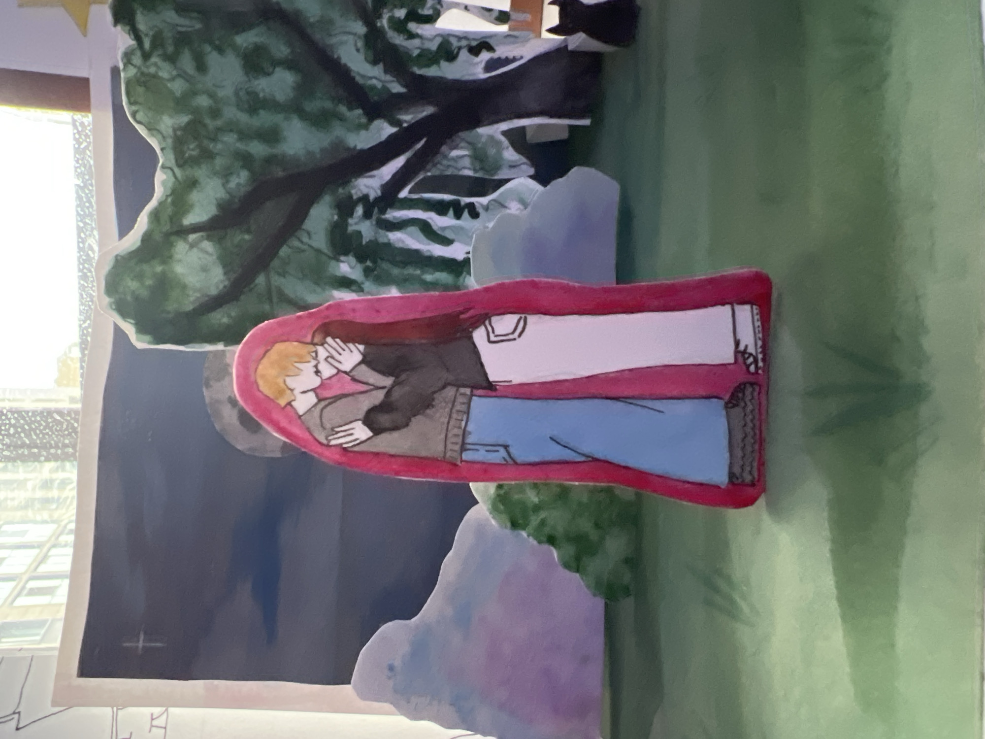 The image displays a handcrafted, multi-layered paper art piece or diorama, oriented vertically. The central element is a cartoon-style illustration of a couple embracing intimately, enclosed within a prominent red, somewhat heart-shaped or oval border. The man, with blonde hair, is on the left, wearing a dark jacket, grey sweater, and blue jeans. The woman, with long dark hair, is on the right, dressed in a white top and pants. Both figures have their eyes closed, conveying a moment of tenderness and affection.
Behind the embracing couple, a nighttime outdoor scene unfolds with layered paper cutouts. Large, stylized green trees with dark branches are visible in the upper sections, suggesting a wooded or park setting. Below the trees, light blue and purple shapes likely represent bushes or clouds. The background is a deep, dark blue, indicating a night sky, where a large, grey full moon is positioned just above the couple. A small white cross symbol is drawn on the dark background to the left of the moon.
The entire setup appears to be displayed on a surface, with a window visible on the far left, reflecting indistinct city buildings. The art piece evokes a romantic and serene atmosphere, likely set during the evening or night. No specific location cues for Saint Petersburg, Russia, are visible within the artwork itself. There is no discernible text apart from the small cross shape on the dark background.