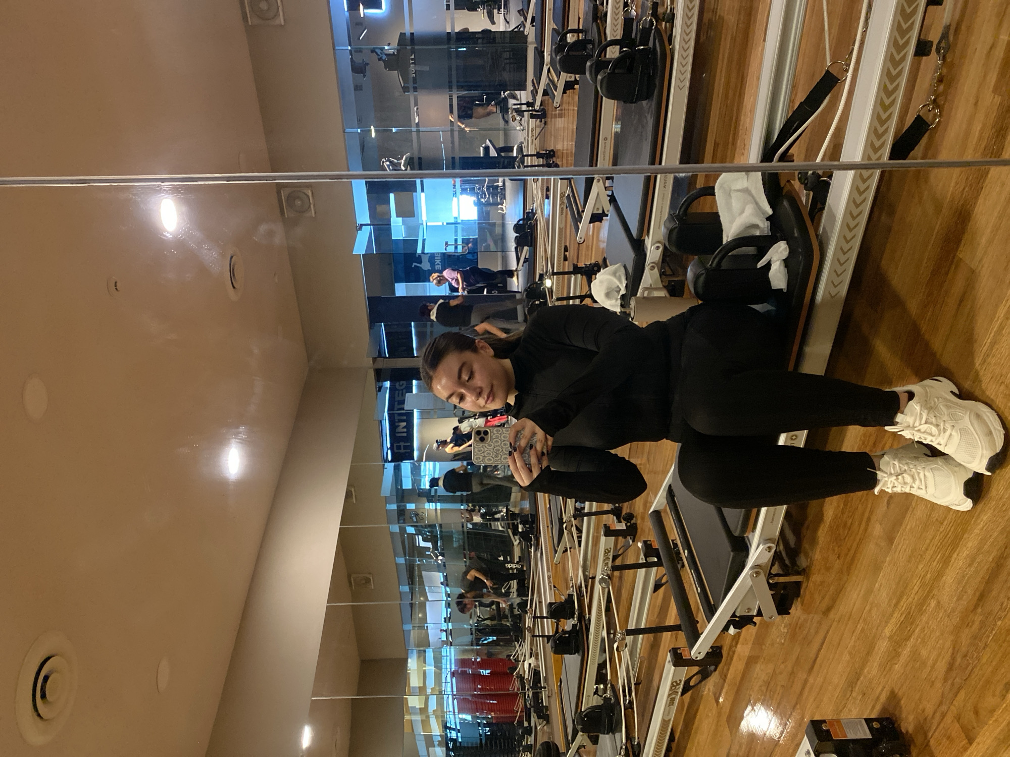 The image shows a young woman sitting on a Pilates reformer machine in what appears to be a gym or fitness studio. The photo is taken in front of a large mirror, capturing her reflection as she holds a smartphone, seemingly taking a selfie. She is wearing black athletic leggings, a long-sleeved black top with thumbholes, and white sneakers. Her dark hair is pulled back, and she has a relaxed expression, looking down towards her phone.
The setting features a light-colored wooden floor and large wall mirrors that reflect the spacious gym. In the reflection, multiple other Pilates reformers or similar exercise machines are visible, arranged in rows. Several other individuals can also be seen in the background reflection, engaged in exercise. The ceiling, also visible in the reflection and directly above, has recessed lighting fixtures and circular air vents. A white towel is draped over a part of the Pilates reformer she is sitting on.
Visible text in the reflection includes "INTEG" on a blue sign and possibly "worth" or similar text on the side of one of the machines. The brand "adidas" is also discernible on a person's shirt in the background reflection. The woman's smartphone has a light-colored, patterned case.