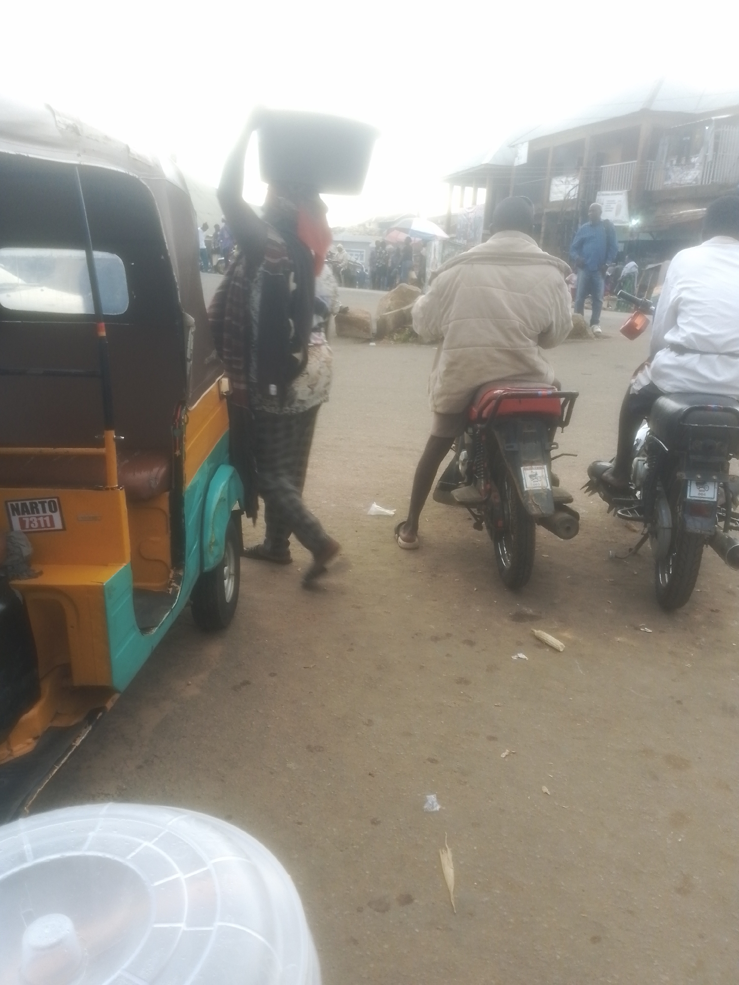 In Gwong, Nigeria, a bright, overcast day unfolds in what appears to be a bustling marketplace or roadside. The scene is dominated by a yellow and teal auto-rickshaw, its door open, parked on a dusty, paved surface. To the left of the auto-rickshaw, a person stands, balancing a large, dark rectangular container on their head. They are dressed in a patterned shirt and pants, with a checkered scarf draped over their shoulders.
Further into the scene, two individuals are seated on motorcycles. The person on the left is wearing a light-colored, quilted jacket and dark shorts, facing away from the camera. The motorcycle they are on is red and black. To their right, another person on a black motorcycle is also facing away, wearing a white long-sleeved shirt. The motorcycles are positioned one behind the other.
In the background, a two-story building with balconies and a corrugated metal roof is visible, suggesting a commercial or residential area. Other people can be seen in the distance, some near stalls or shops, implying a lively gathering. There are some scattered rocks and debris on the ground, including what looks like a piece of dried corn.
The visible text on the auto-rickshaw reads "NARTO 7311." The overall atmosphere seems active and functional, typical of a roadside commerce area in Nigeria. The lighting suggests it is daytime, with the overcast sky creating diffused light.