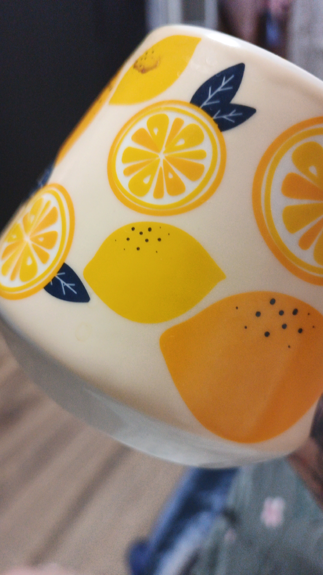 The image shows a close-up of a ceramic mug decorated with yellow lemons and orange slices. The mug is light-colored, possibly cream or off-white, and has a glossy finish. The designs are simple and stylized, with dark blue leaves accenting some of the fruit. One large yellow lemon is prominent in the foreground, with several black dots representing seeds. To the left of this lemon is a slice of an orange, and above it, another slice of an orange and a whole lemon. The background is blurred, but appears to be an indoor setting with a wooden surface below and a dark, possibly black, background behind the mug. There is a hint of a person's clothing, appearing to be denim blue, in the lower right portion of the image. The overall impression is of a bright, cheerful kitchen or dining setting. The mug itself is positioned on an angle, obscuring a full view of its surface. No information is available about the location in the city of Drezna, Russia.