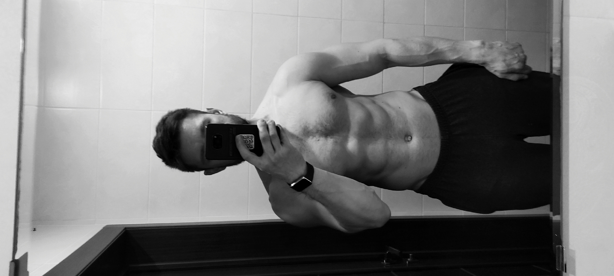A shirtless man takes a mirror selfie in a bathroom with white tiled walls. He is holding a smartphone in his left hand, obscuring part of his face. He wears dark shorts and a smartwatch on his left wrist. A dark-colored doorframe is visible below the mirror. The image is in black and white. There is no information suggesting the location is Fryazino, Russia.