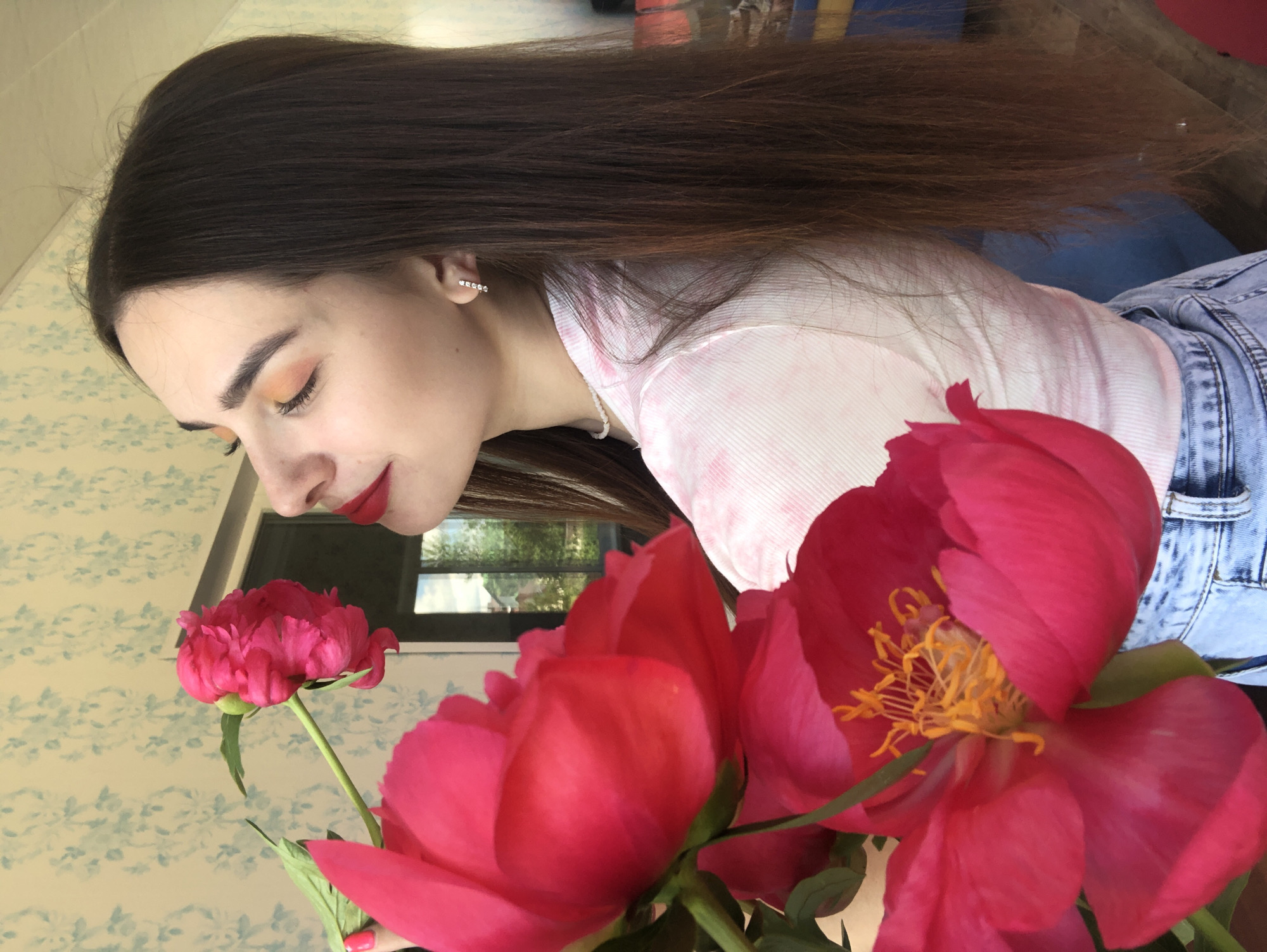 A young woman with long, dark brown hair and fair skin is standing indoors, near a window. She is wearing a light pink ribbed t-shirt and light blue jeans. She is looking towards the right side of the frame, with her eyes closed, and her head tilted slightly back as if smelling flowers. In the foreground, on the left side of the image, are several large, bright pink peonies. The woman has on red lipstick, orange eyeshadow, and a small pearl necklace. She is also wearing a square, diamond stud earring in her right ear. Behind her, a window shows a blurred view of trees and buildings, suggesting an outdoor scene beyond. The wall behind the woman has a light blue floral wallpaper pattern. The lighting appears to be natural, coming from the window. The overall scene conveys a sense of peaceful enjoyment of the flowers. The specific location in Noviy, Russia, is not discernible from the visual information in the image.