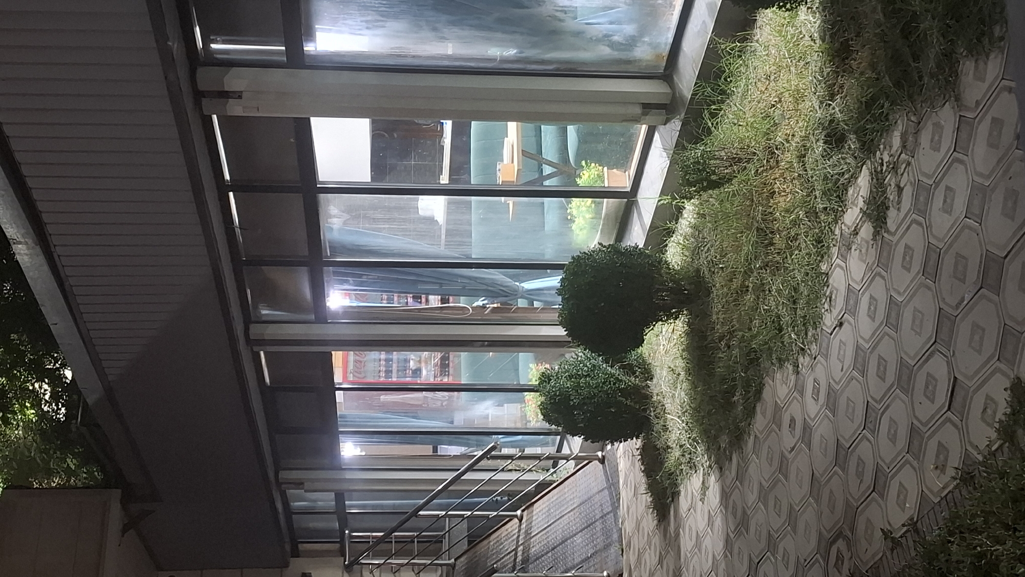 This is a medium shot of an exterior passageway with a modern architectural design. The scene is illuminated by artificial lights, suggesting it could be evening or night. The overall atmosphere appears tranquil and somewhat moody.
The primary subjects are architectural elements and landscaping. A series of large glass windows form a significant part of the structure, allowing glimpses into an interior space that appears to be a cafe or lounge, with tables and seating visible. The windows are framed by dark metal, and a portion of a corrugated metal roof extends over the passageway.
To the right of the passageway, a section of meticulously landscaped area features a grassy bank that cascades over a textured concrete pathway. The pathway is paved with hexagonal tiles, each containing a smaller octagonal detail in a lighter shade. Two perfectly spherical topiary bushes stand prominently in the grassy area.
The left side of the image shows a modern metal staircase with a sleek railing, leading downwards into the structure. The building itself has a clean, contemporary aesthetic. The lighting creates a contrast between the illuminated areas and the darker shadows cast by the roof. There is no visible text in the image. The location context of Shymkent, Kazakhstan, does not provide specific visual cues within the image itself, but frames the setting as a modern urban environment in that region. No people are visible, and the overall impression is one of a quiet, well-maintained public or commercial space.
