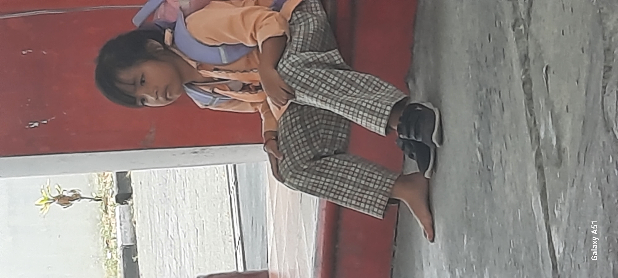The image shows a young girl sitting on a red step, wearing an orange shirt, checkered pants, a blue backpack, and dark shoes. She is barefoot on her left foot. In the background, there is a textured red wall and a small plant. The scene appears to be outdoors, possibly in a public area. The lower right corner of the image displays the text "Galaxy A51". The location is identified as Ploso, Indonesia.