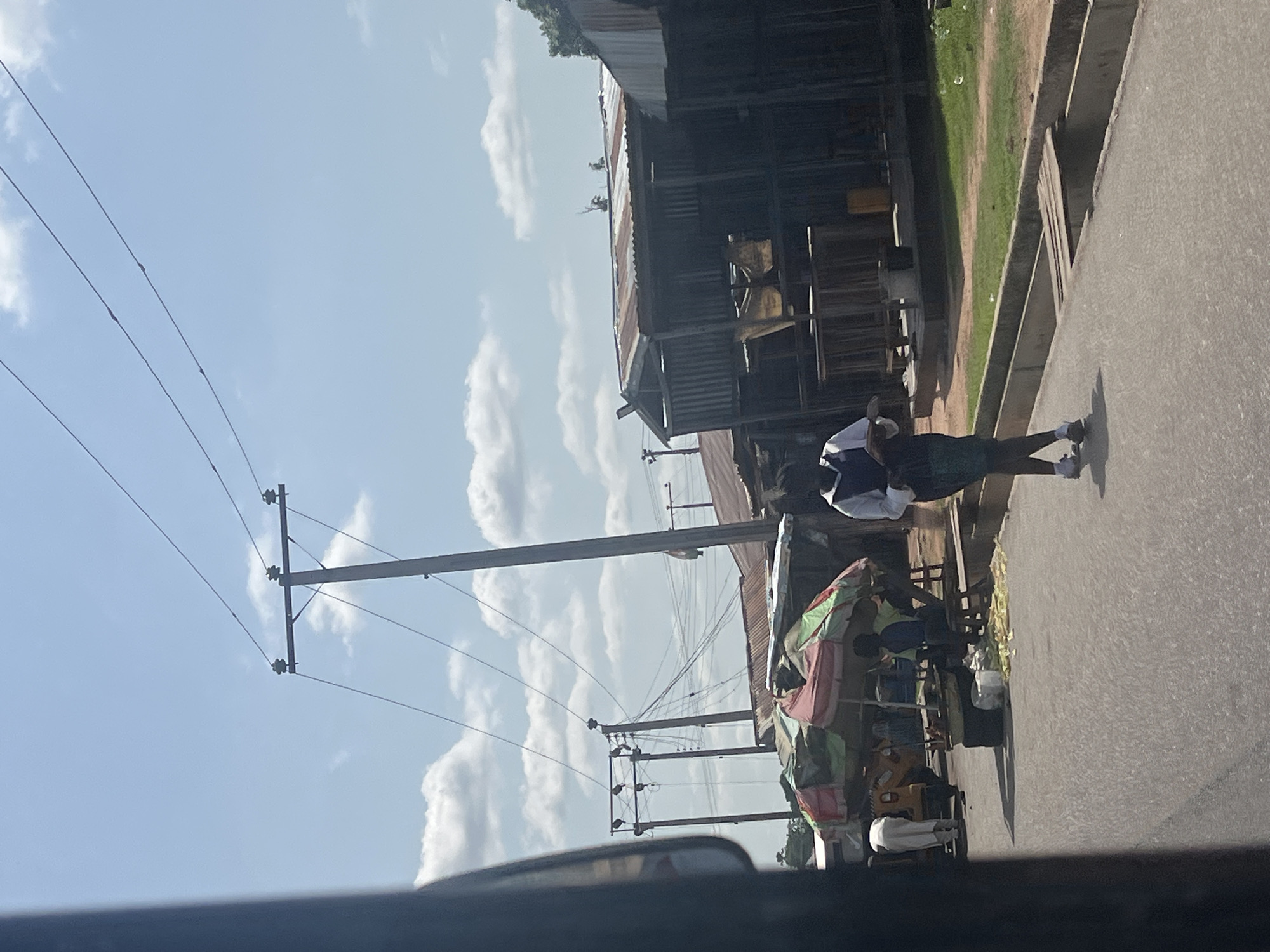 In Jalingo, Nigeria, on a sunny day with scattered clouds, a street scene unfolds. A young woman, wearing a white shirt with a dark vest and a patterned skirt, walks along a paved road. She is holding a tablet or book in her hands. To her left, a market stall is set up under a multi-colored umbrella, with several people gathered. A yellow auto-rickshaw is parked nearby. Behind the stall, wooden structures with corrugated metal roofs form shops. Utility poles with electrical wires crisscross the sky above the scene, indicating the presence of infrastructure within the urban environment. The road is lined with a concrete drainage channel.