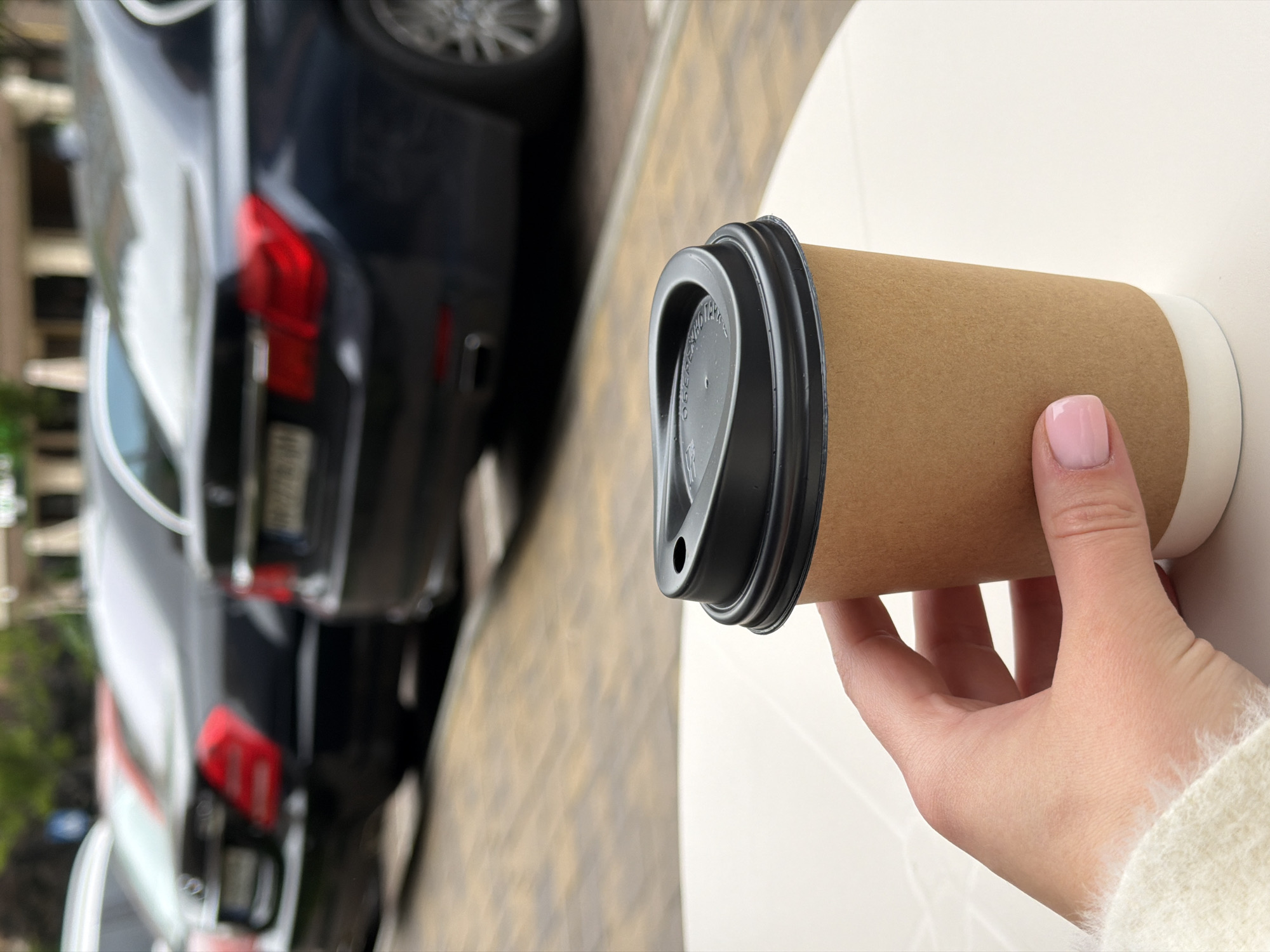 A close-up, tilted shot captures a person's hand holding a disposable coffee cup. The hand, with light pink nail polish on the visible thumb and fingers, is wearing a light-colored, possibly fuzzy or knit, sweater. The coffee cup is made of plain brown kraft paper with a black plastic lid. Partially visible text on the lid is largely illegible due to blur and angle.
The cup is held over a white, smooth surface, likely a table or ledge, which occupies the right side of the frame. In the blurry background on the left, a dark-colored car, possibly a sedan, is parked on a patterned paved surface, which could be asphalt or cobblestones. The car's red taillights are visible. The scene appears to be outdoors during the daytime, with natural lighting. The overall setting suggests an urban or suburban outdoor area in Sofiivska Borshchahivka, Ukraine.
