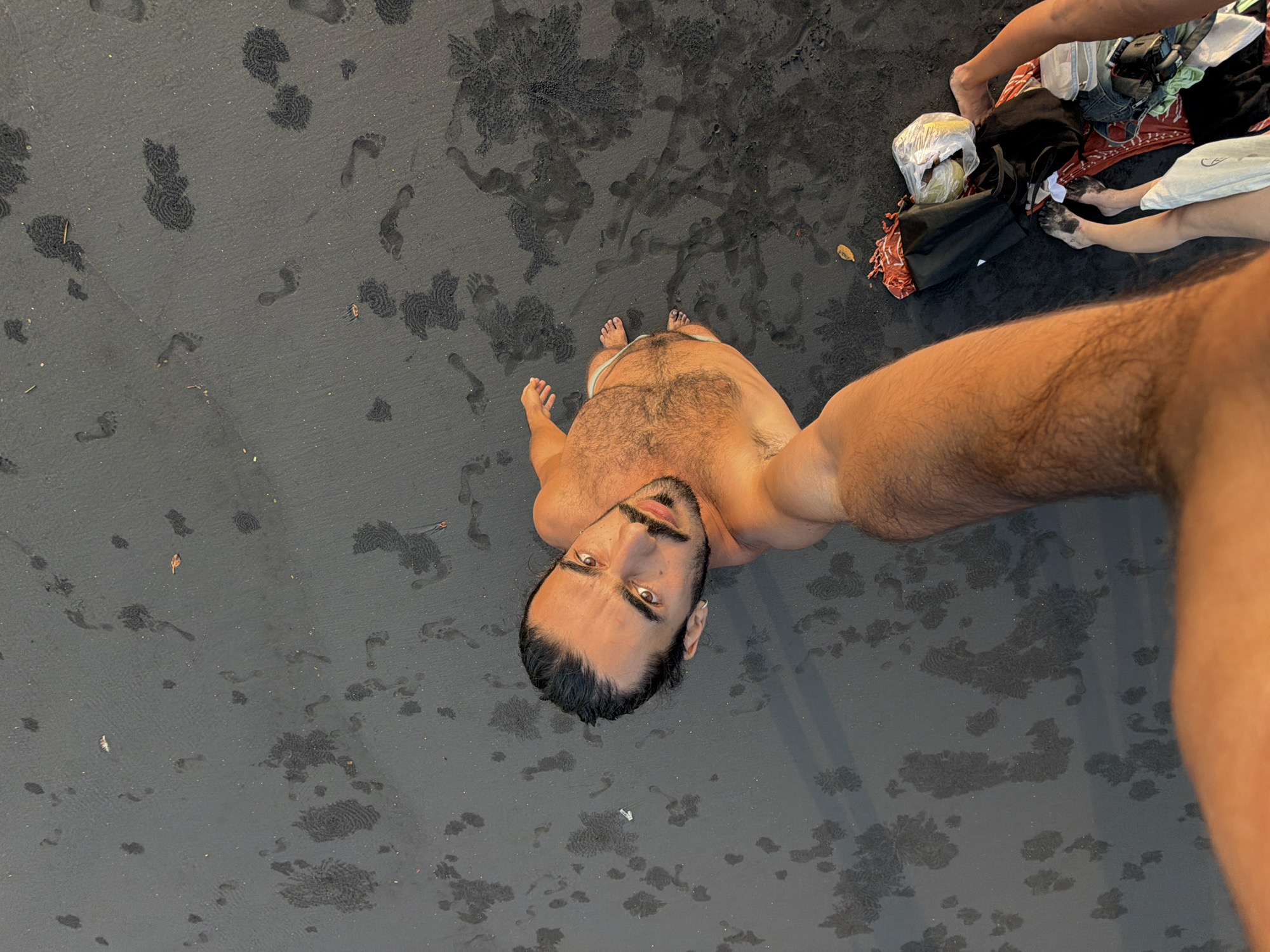 A high-angle, full shot captures a man taking a selfie on a dark, wet sand beach. The man, with dark hair pulled back and a beard, is shirtless and wearing light-colored swim trunks. His right arm is extended towards the camera, holding it. The sand is marked with numerous footprints and what appear to be areas where water has pooled and dried, creating intricate patterns. To the lower right of the frame, a pile of clothing and bags lies on the sand, with a pair of legs emerging from the pile. The scene is illuminated by warm, golden-hour light. The location is identified as Selemadeg Timur, Indonesia.