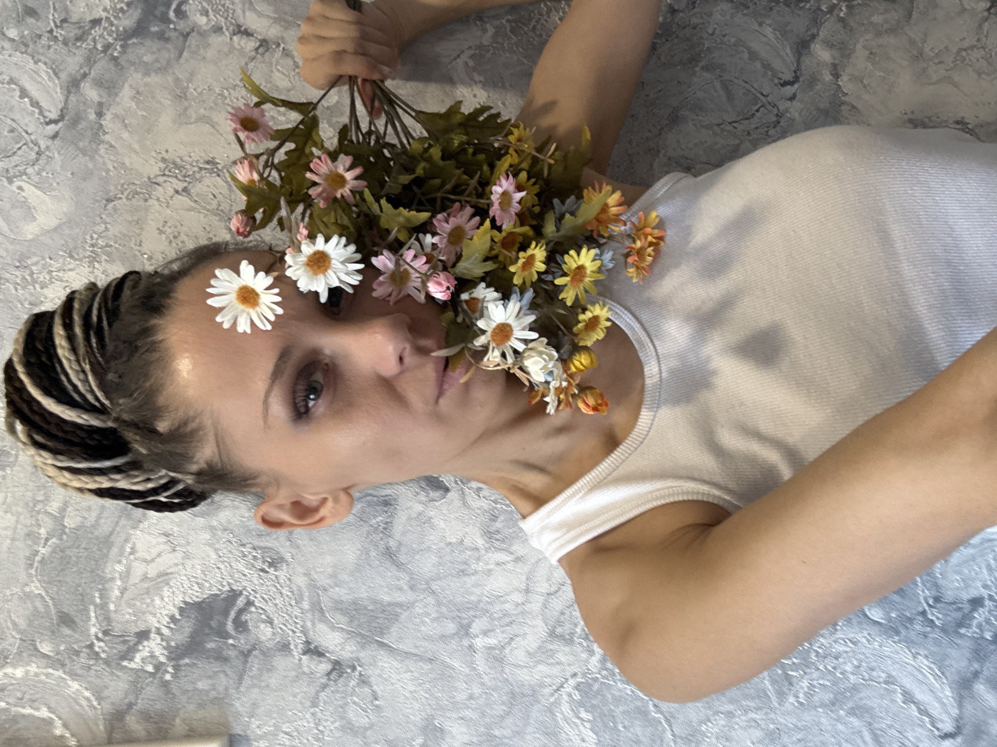 A person with braided hair and a white tank top is lying down on a grey textured surface, possibly a bed or wall, while holding a bouquet of colorful flowers near their face. The flowers consist of white, pink, orange, and yellow daisy-like blooms. The person's expression is neutral, and they appear to be looking at the camera.
There is no information about Rusanovo, Russia in this image.