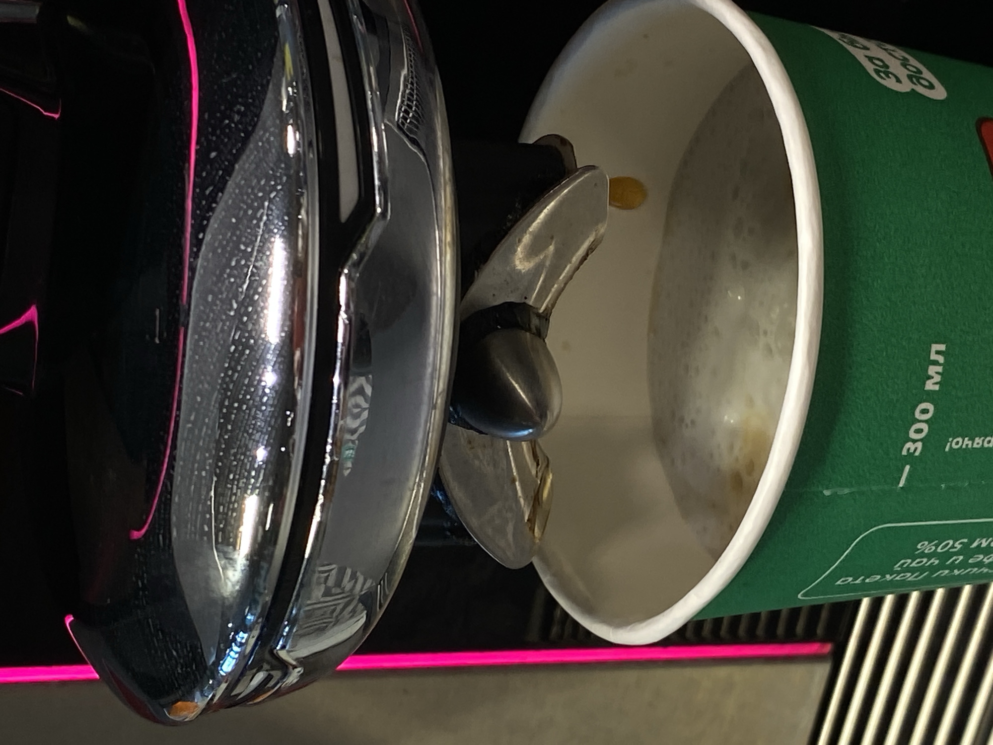 Here's a matter-of-fact description of the image:
The image shows a close-up of a coffee machine dispensing coffee into a green and white paper cup. The coffee has a foamy appearance. The coffee machine is black and chrome, with pink neon lighting accents. Text in Russian is visible on the side of the cup. The scene appears to be indoors.