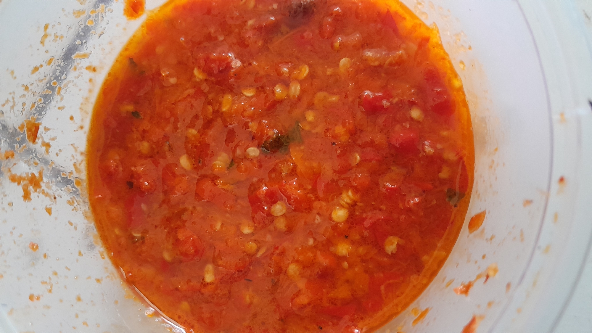 A close-up, overhead view of a bowl of vibrant red chili sauce. The sauce is a thick, textured blend, with visible chili seeds and small pieces of chili flesh suspended in a liquid base that appears to have a slight sheen of oil. The sauce fills the majority of the frame, with the translucent plastic of the bowl visible around the edges. The bowl itself appears slightly smudged with traces of the chili sauce on its interior walls. The background is out of focus, providing no discernible environmental context. This image was taken in the city of Bandung City, Indonesia.