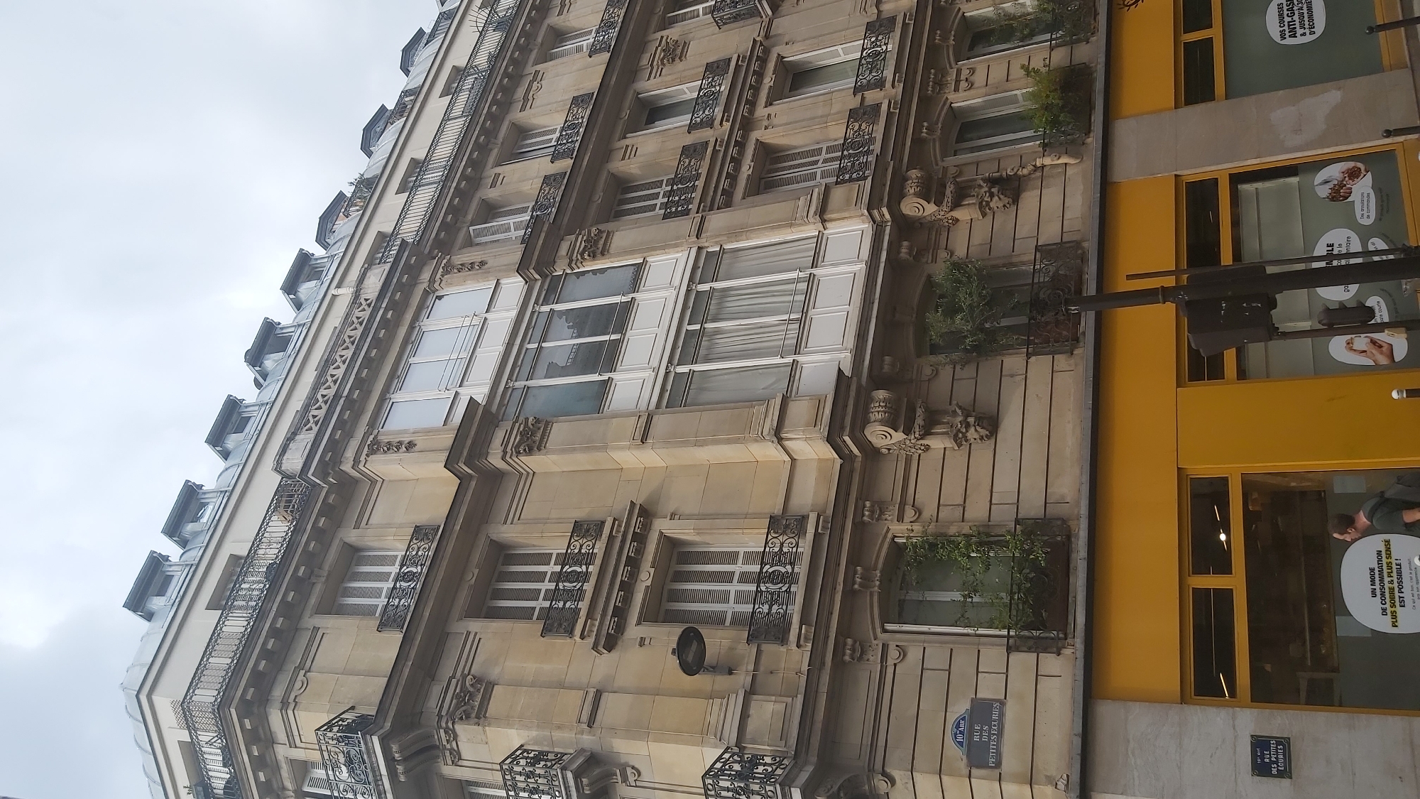 A low-angle, full shot captures the facade of a building in Paris, France. The building is constructed from light brown stone with decorative carvings and features multiple floors with balconies and windows. The balconies have ornate black iron railings. The upper floors have mansard roofs with dormer windows. A yellow awning runs across the bottom of the building, above which are shop windows. A street sign that reads "10e Arr. Rue des Petites Ecuries" is visible on the left side of the building. A man is walking by on the sidewalk in front of the shop.