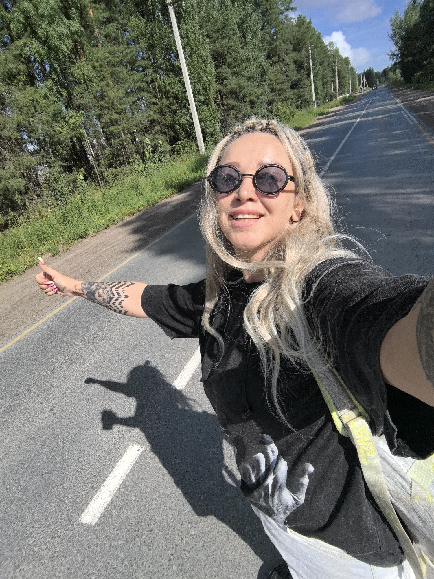 A blonde woman with braided hair and sunglasses is standing on the side of a road, hitchhiking. She is wearing a black t-shirt with a graphic print and white pants. Her left arm is extended with her thumb up, and she has tattoos on her arm. The road appears to be in a rural area, with trees lining the sides. The sky is blue with some clouds. The image is a selfie taken by the woman. The location is identified as Chaikovsky, Russia, based on metadata or external information not present in the image itself.