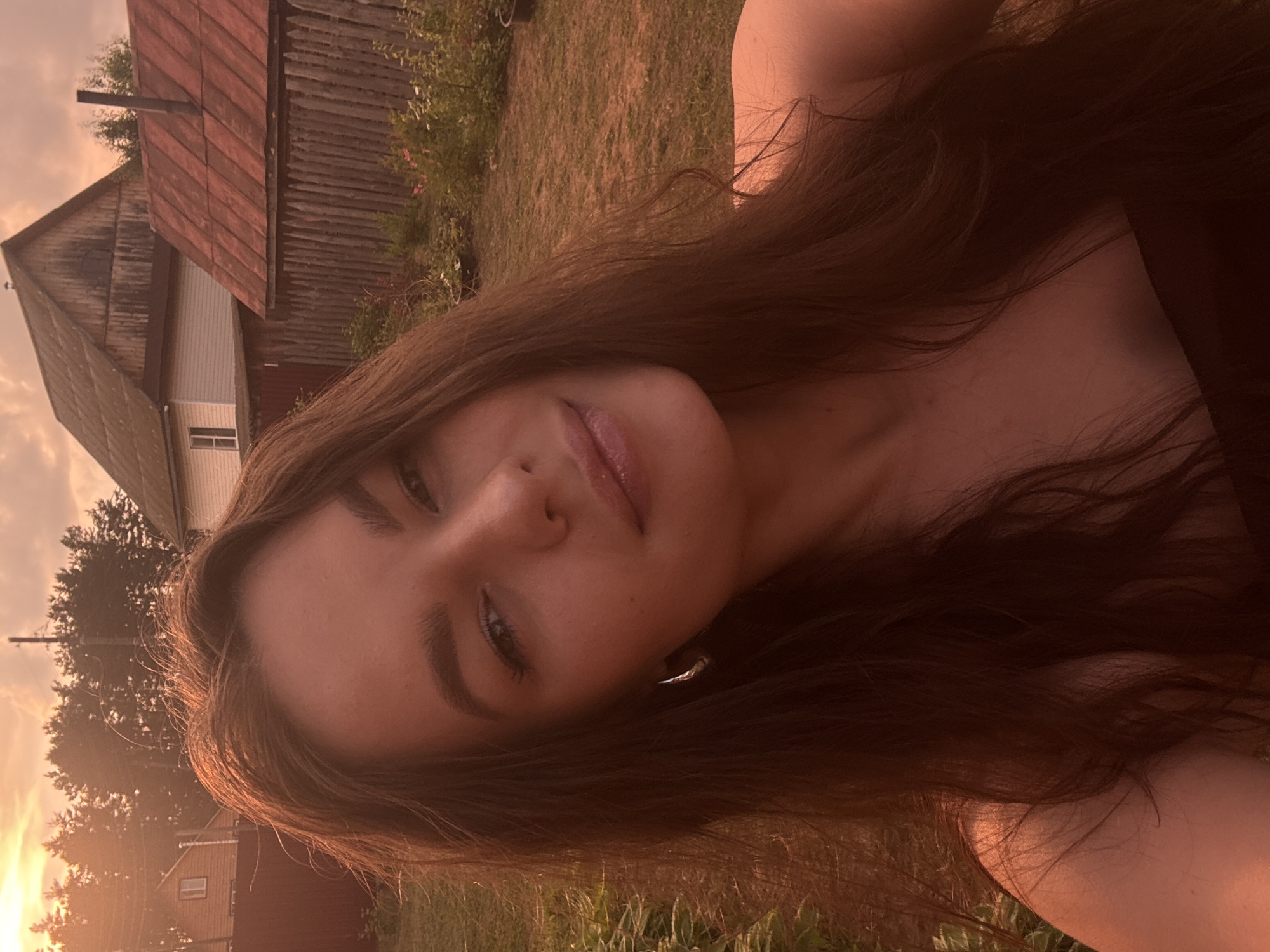 The image is a close-up, outdoor selfie taken during sunset in Rusalevka, Russia. A young woman with long, brown hair is the primary subject, positioned in the foreground and looking towards the camera. Her face is illuminated by the warm, golden light of the setting sun, creating a soft glow. She is wearing a hoop earring on her left ear. In the background, traditional wooden houses and fences are visible, characteristic of a rural Russian village. Trees line the horizon, and the sky is a warm gradient of orange and pink hues, indicative of a sunset. The ground appears to be dirt or grass, with some sparse vegetation. The overall scene conveys a sense of peacefulness and natural beauty.