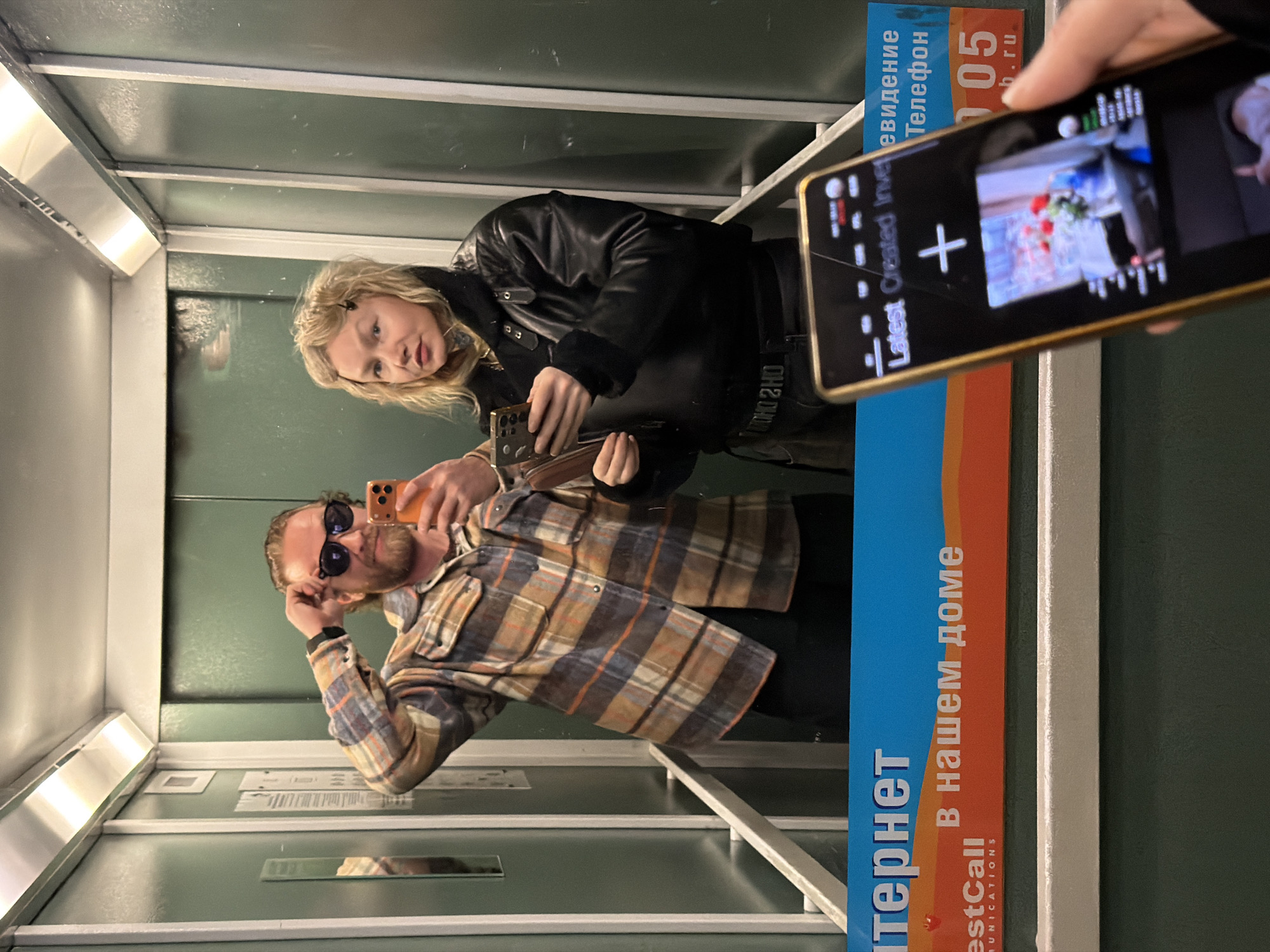 The image depicts two individuals taking a selfie in an elevator. A man with a beard and sunglasses is holding a phone, presumably taking the photo. A woman is standing beside him, also holding a phone. Another phone is held by a third person in the foreground, displaying a recording with visual elements. The elevator's interior is visible, including advertising panels with Russian text, identifying the location as Saint Petersburg, Russia. The text on the panel says, "Internet in our home." The elevator has green walls.