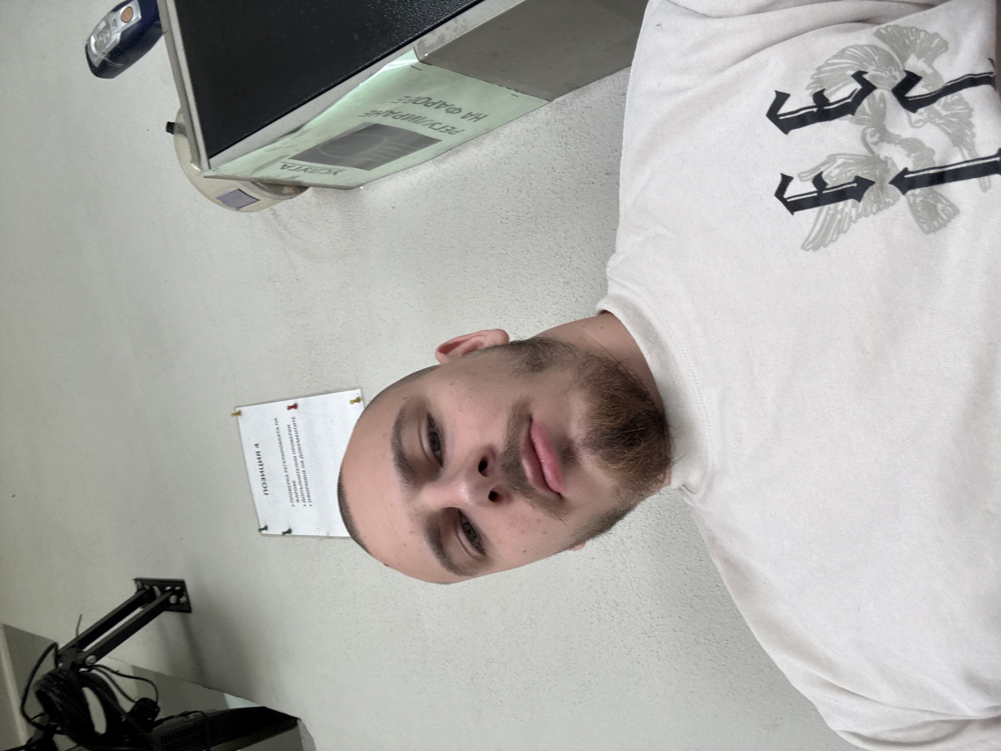 The image shows a man with a beard, wearing a white long-sleeved shirt with a graphic print, in an indoor setting. To his left is a computer monitor on a stand. A sign is posted on the wall that is written in Bulgarian, mentioning "проверка регулировката на фарове" which relates to checking headlight alignment. Another sign reads “Position 4” and refers to items checked and documented. The context suggests a vehicle inspection or repair facility in Stara Zagora, Bulgaria.