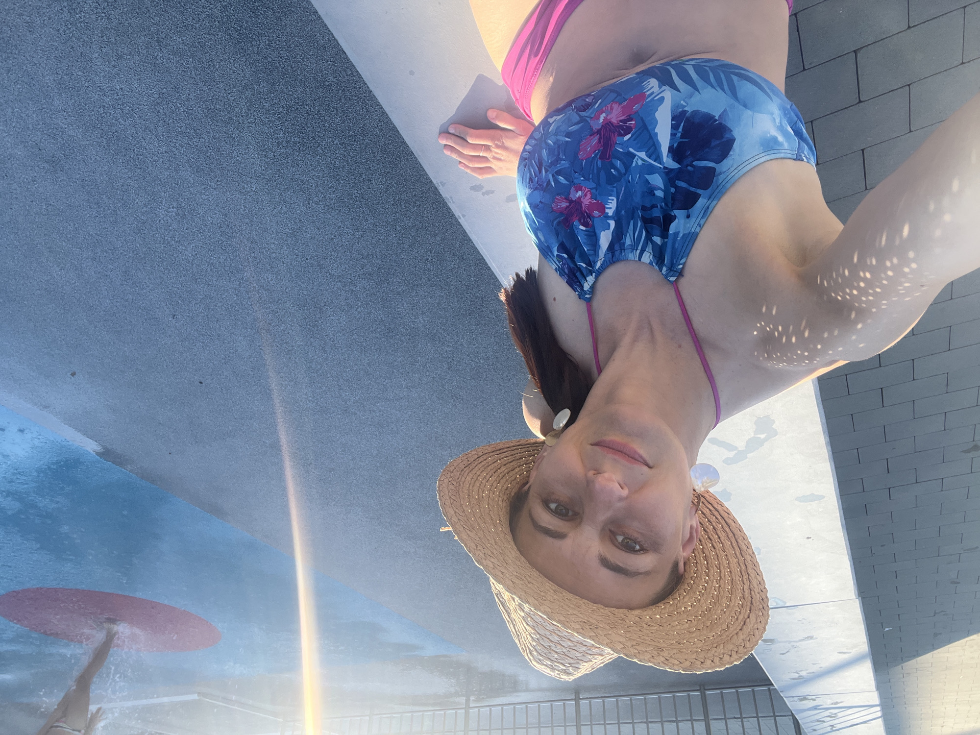 The image shows a woman taking a selfie at a pool. She is wearing a blue and pink floral bikini and a straw hat. The woman is smiling and looking at the camera. The setting appears to be a sunny day at a swimming pool, possibly a water park, with a textured grey surface and a blue pool. Light is reflecting off the water, creating dappled patterns on her skin. In the background, a section of the pool is visible with a red disc on the water, and another person can be seen in the distance. The overall mood is relaxed and happy, suggesting a day of leisure and enjoyment.